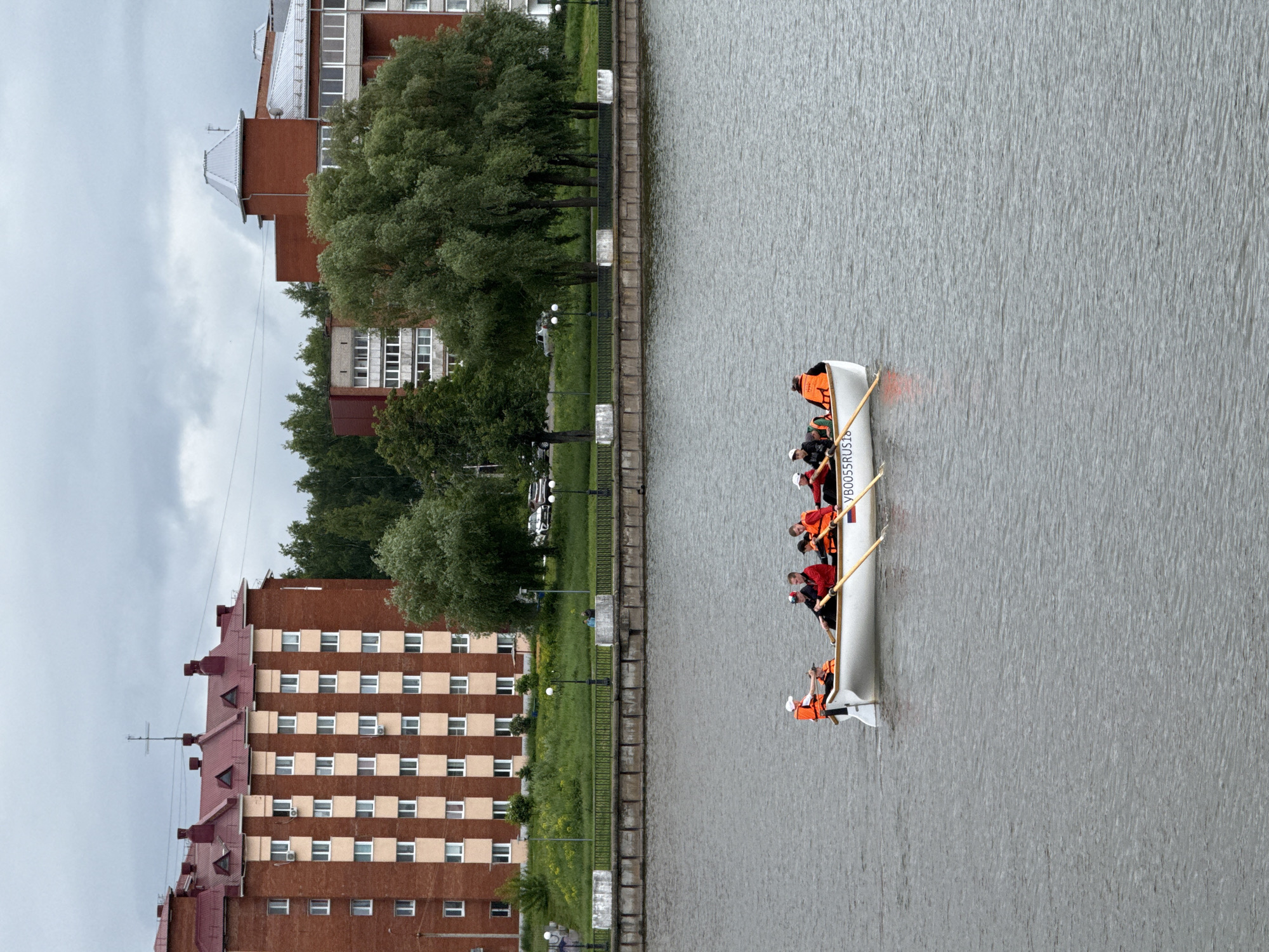 This image captures a wide view of a rowing scene on a river in Votkinsk, Russia, under an overcast sky. In the foreground, a white, long rowing boat occupies the center of the frame, moving left to right across the gray, slightly rippled water. Seven individuals are in the boat. Six are seated and engaged in rowing, wearing either orange or red life jackets and holding wooden oars. One person is standing at the stern, possibly a coxswain, also wearing an orange life jacket and a white cap. The side of the boat displays the registration number "VB0055RUS18" and a small flag resembling the Russian tricolor.
The midground features the river's embankment. A concrete retaining wall lines the shore, topped by a green grassy slope. Above the slope, a paved path is visible, intermittently lit by black lampposts. A dark metal fence runs parallel to the path, separating it from the grassy area and sparse trees. In the distance on the embankment, several parked cars are discernible.
The background is dominated by urban architecture and abundant greenery. To the left, a multi-story red brick residential building stands prominently, featuring a dark red gabled roof with several dormer windows and a cross-like antenna on its peak. The building's facade is characterized by repetitive rows of windows, some with light-colored frames, and vertical sections of a lighter hue between window columns. Several air conditioning units are attached to the exterior walls. To the right, behind a dense cluster of trees, another red brick building complex is visible, distinguished by a lighter gray, possibly metallic, roof and a prominent conical or pyramidal roof structure on one section. Large trees with lush green foliage are interspersed across the background, obscuring parts of the structures and creating a natural buffer between the river and the city. The sky is uniformly gray, indicating a cloudy day.