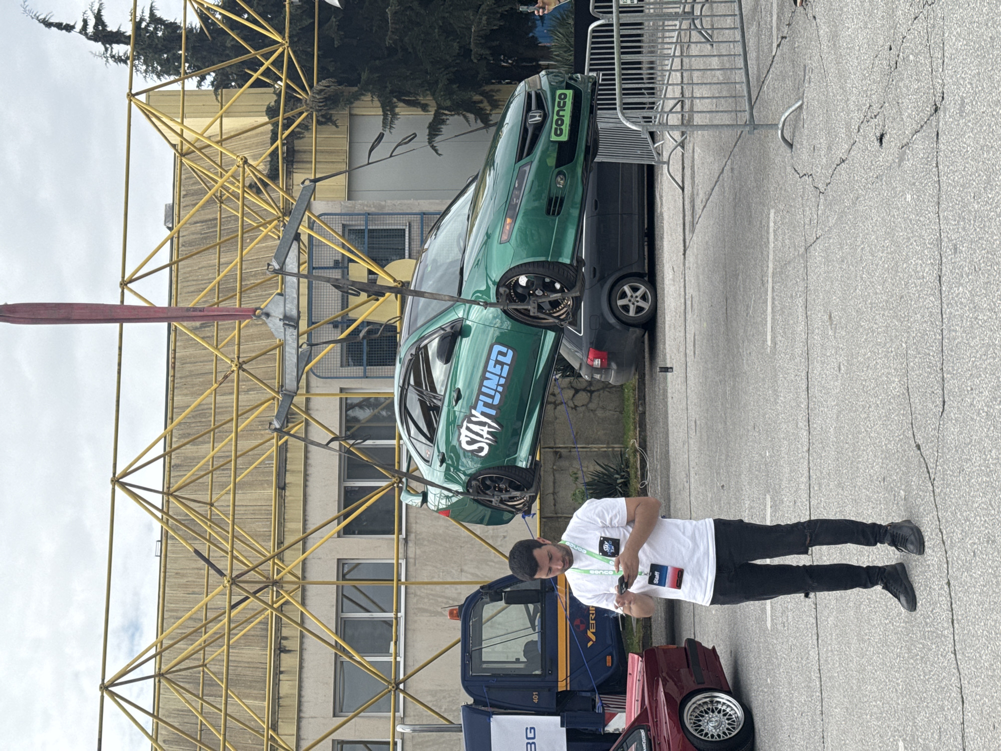 The image captures an outdoor scene in Plovdiv, Bulgaria. A green Honda car with "STAY TUNED" graphics is elevated by a yellow scaffolding structure. Beneath the elevated car is a dark blue tow truck. A man wearing a white t-shirt, black jeans, and black shoes is standing in the foreground, looking at his smartphone. He has a "Staff" badge and a lanyard around his neck. A maroon car is partially visible on the right side. The background features a building with multiple windows and a metal fence. The ground appears to be asphalt. The weather seems overcast.