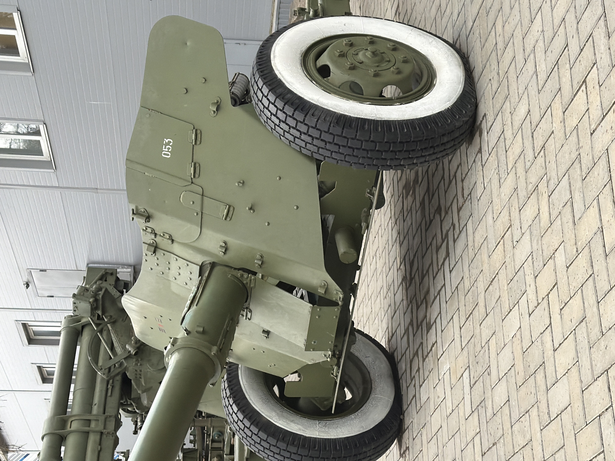The image shows a 57mm anti-tank gun, likely a ZiS-2 or similar model, displayed at the "Enchanted Smithy" Museum in Votkinsk, Russia. The gun is painted in a matte green color and features white-walled tires on its wheels. The number "053" is stenciled on one of the gun shields. The background includes a building with horizontal siding and rectangular windows. The ground is paved with light-colored bricks laid in a herringbone pattern. The photograph is taken from a low angle, looking upwards at the artillery piece.