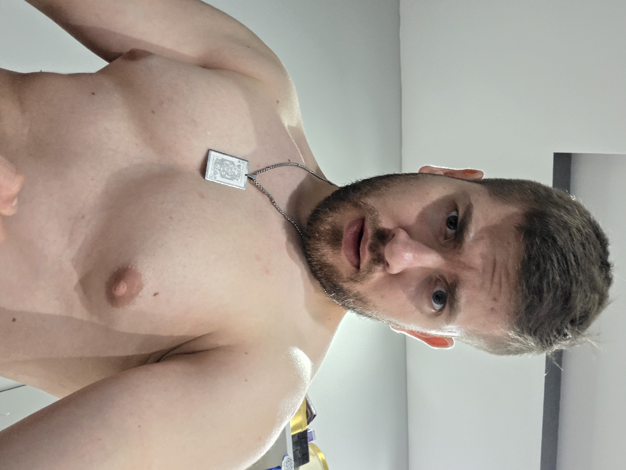 A Caucasian man with short brown hair and a beard is positioned shirtless indoors against a white wall with a black vertical accent. He is wearing a silver chain necklace with a rectangular pendant. The lighting is bright.