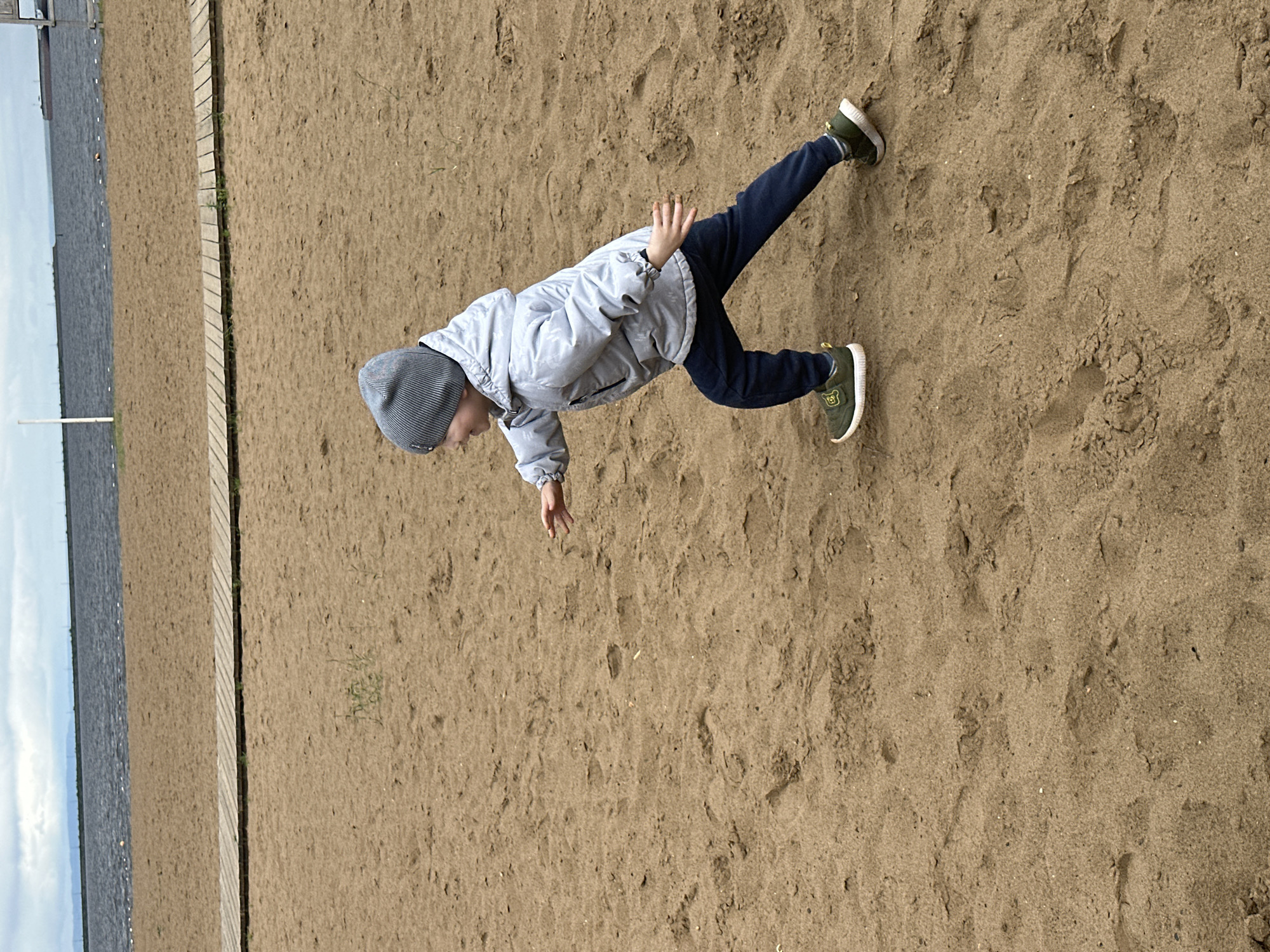 The image depicts a young child in motion on a sandy beach in Chaikovsky, Russia. The child, appearing to be a toddler or preschooler, is captured mid-stride, moving from left to right across the frame. They wear a grey ribbed knit beanie, a light grey or silver hooded jacket with a faint patterned texture, dark blue trousers, and olive green athletic shoes with white soles and yellow detailing. Their mouth is open in a smile, and their arms are bent and extended forward with open palms, suggesting a running, reaching, or playful gesture.
The ground consists of light brown, fine-grained sand, showing multiple footprints and sparse tufts of light green vegetation. A light-colored wooden boardwalk, constructed from horizontal planks, runs parallel to the water's edge across the mid-ground. Beyond the boardwalk is a dark blue-grey body of water, presumably a large lake or river, with visible surface choppiness and several white buoys. A distant, low-lying landmass or industrial structures are visible on the far horizon across the water. A large, dark-hulled ship is faintly discernible on the far right horizon over the water. The sky is overcast with light grey clouds, indicating diffuse lighting conditions. A white vertical pole is present on the left side of the frame, near the water line.