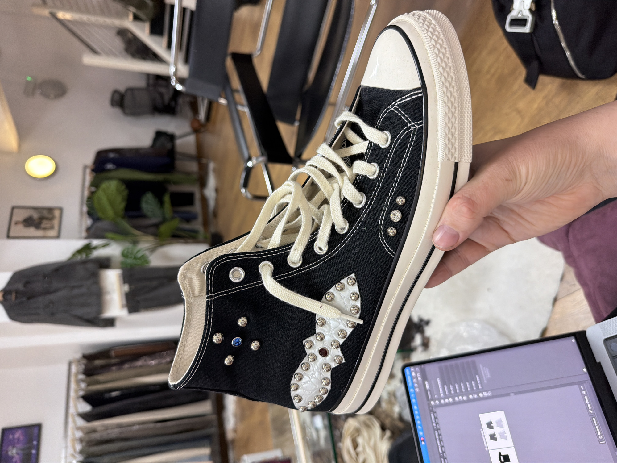 The scene depicts the interior of a fashion boutique or custom workshop in London, United Kingdom. A person's hand, exhibiting fair skin and short fingernails, holds up a single black high-top canvas sneaker for display. The sneaker features white stitching, cream-colored laces, a white rubber toe cap, and an off-white rubber sole with a textured sidewall. It is customized with multiple silver dome studs and rhinestones: a single blue rhinestone near the top lace eyelet, a larger clear rhinestone on the lower lateral side, and a smaller clear one below it. A white, textured material appliqué, shaped like a stylized wing or abstract design, is affixed to the lateral side, adorned with numerous small silver dome studs and a central red rhinestone. In the immediate foreground, a silver MacBook laptop is open, displaying graphic design software featuring product mock-ups, resting on a purple fabric item. A white, possibly faux fur, rug is on the light-colored wooden floor beneath the laptop. A black bag with a silver buckle is visible on the far right. The background environment includes a clothing rack holding various garments in muted tones, a large green potted plant, black modern chairs with metal frames, a white wall with a circular yellow light fixture, a framed portrait, and a modern staircase with metal railings on the right. No other individuals are visibly present beyond the holding hand.
