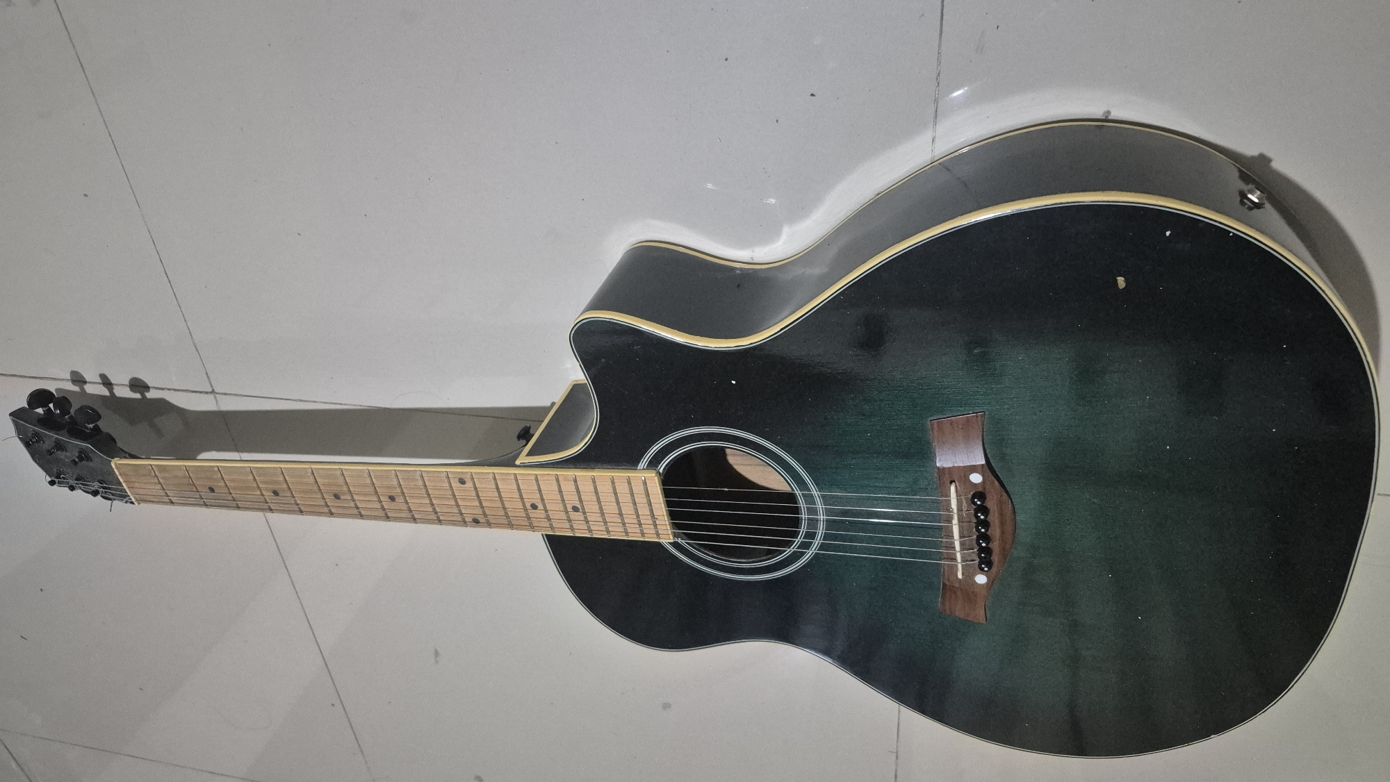 The image shows an acoustic guitar with a dark green to black gradient finish, leaning against a light-colored tiled wall. The guitar has a cutaway body and a wooden bridge with six black bridge pins. The fretboard is light-colored wood, and the headstock has black tuning pegs. A shadow of the guitar is cast on the wall. The scene appears to be indoors, possibly in a home or studio setting, and there is no indication of it being located in the city of Bojonegoro, Indonesia. The image is a close-up, eye-level shot of the guitar.