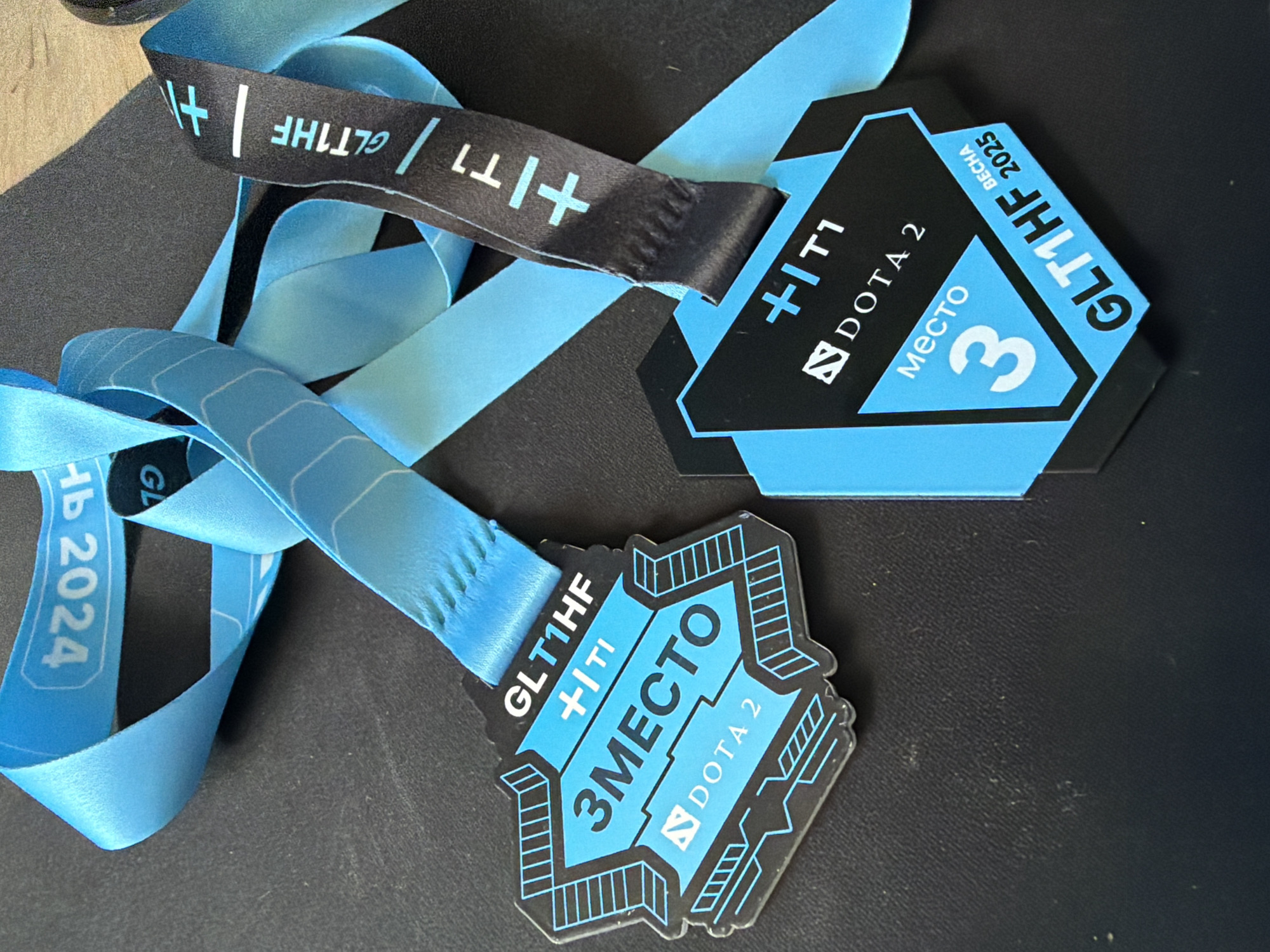 The image displays two esports medals and their associated ribbons, resting on a dark, possibly black, textured surface. Both medals and ribbons feature a distinctive black and light blue color scheme with a modern, geometric design.
One medal, octagonal in shape, prominently features the text "GLT1HF", "+T1", and the "DOTA 2" logo. Below these, "МЕСТО 3" (meaning "3rd place" in Russian) is displayed, with "ВЕСНА 2025" (meaning "Spring 2025") printed vertically along its right edge. This medal is attached to a light blue ribbon.
The second medal has a more complex, angular, and outlined shape. It also shows "GLT1HF", "+T1", and "ЗМЕСТО" (3rd place) centrally, with the "DOTA 2" logo underneath. This medal is connected to a light blue ribbon.
Visible among the light blue ribbons is also a black ribbon patterned with "GLT1HF" and "+T1" text in white. Another section of a light blue ribbon clearly shows "ВЕСНА 2024" (Spring 2024).
The awards signify a third-place achievement in a DOTA 2 esports tournament, likely sponsored or organized by entities named "GLT1HF" and "+T1". The use of Cyrillic script for "место" and "весна" indicates a Russian-speaking context, consistent with the location of Chaikovsky, Russia. The different years (2024 and 2025) visible on the ribbons and medals suggest either different tournament editions or perhaps a ribbon from an earlier event paired with a newer medal. The overall presentation is a celebration of competitive gaming success.