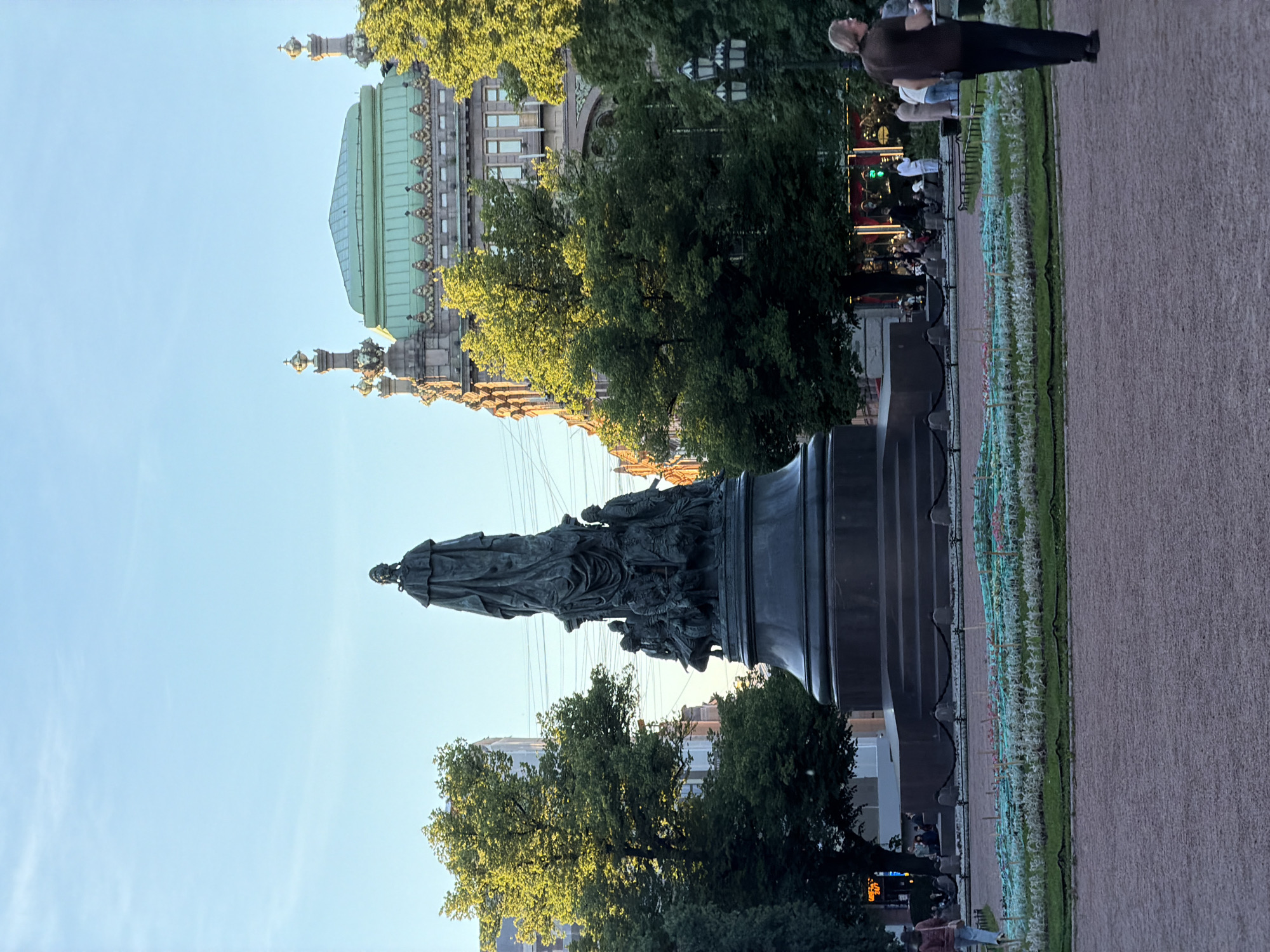 The photograph captures a view of the Monument to Catherine the Great in Nevsky Prospect, Saint Petersburg, Russia. The monument, a large bronze sculpture depicting Catherine II and allegorical figures, stands prominently in the center of the frame. It is situated within a manicured garden area featuring a decorative low fence and flower beds. In the background, several buildings with distinctive architectural features are visible, including one with a green roof and ornate detailing. The scene is bathed in the warm light of what appears to be late afternoon or early evening. Several individuals are present in the park, with some seated near the monument and others walking in the foreground and background. The sky above is clear and blue.