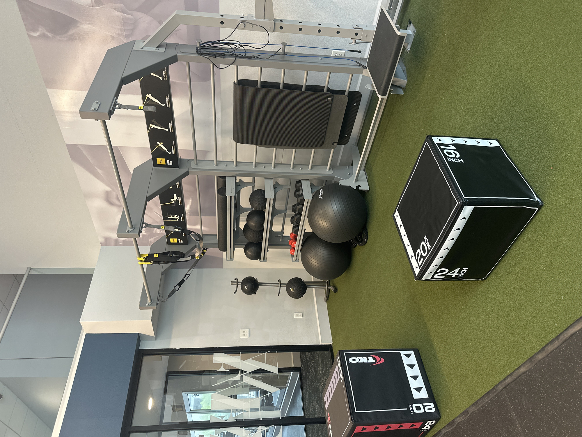 This indoor shot showcases a fitness center, evidenced by the presence of exercise equipment. A weight rack holds various dumbbells and exercise balls. A black and white plyometric box is seen to the right of the rack, while another plyometric box is at the bottom of the image. The location appears to be in Clifton, United States of America. The scene is devoid of people.
