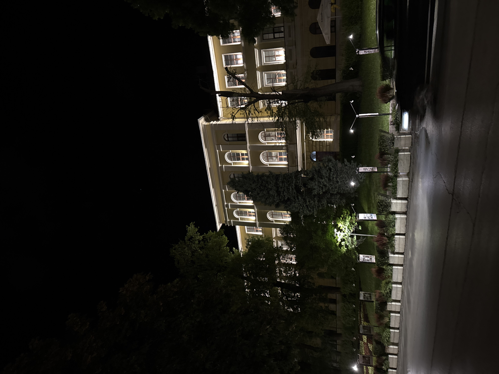 At night, a four-story, yellow-beige building with many windows illuminated is visible. The building's architectural style is classic, with rows of arched windows on the lower floors and rectangular windows above. A large tree stands in front of the building, partially obscuring the view. Street lamps illuminate a row of flower beds and small markers or signs along a sidewalk. The scene appears to be wet or recently rained on, as the pavement reflects the light. The location appears to be in Veliko Tarnovo, Bulgaria, based on the architectural style and general ambiance. The lack of pedestrian activity suggests a quiet or late-night setting.