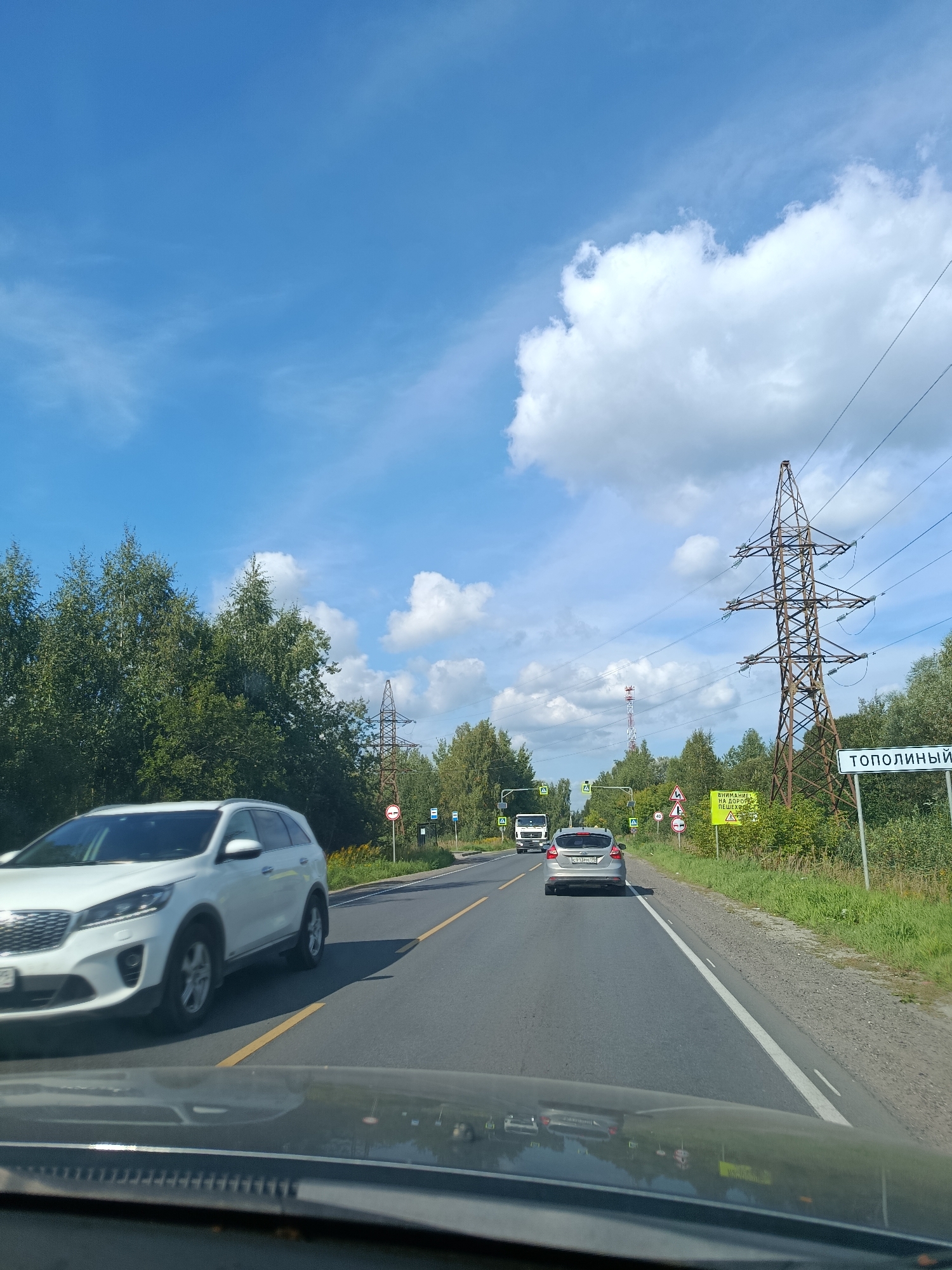 This image captures a view from a vehicle traveling on a two-lane road near the settlement of Тополиный, Russia. The sky is bright blue with scattered white clouds. On the right side of the road, a tall, lattice-style electricity pylon stands prominently. Adjacent to the pylon is a white sign with Cyrillic text that reads "ТОПОЛИНЫЙ." Further down the road on the right, there are smaller road signs, including a yellow warning sign with Cyrillic text and a circular sign with a red border and a diagonal line, likely indicating a restriction. A bus stop sign is also visible.
The road itself is paved and has yellow dividing lines. Several vehicles are on the road: a white SUV is in the left lane, moving away from the camera, and a silver hatchback is in the right lane, also moving away. A white truck is further ahead in the left lane. The left side of the road is lined with dense green trees, characteristic of a rural or semi-rural environment. The overall scene suggests a sunny day on a highway in a forested area.