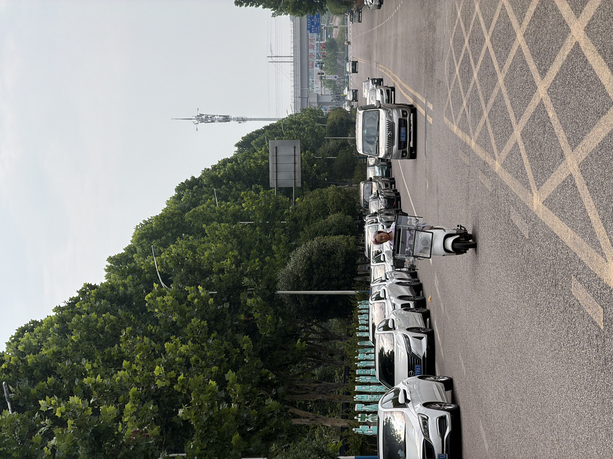 This daytime image captures a street scene in Jiangjin, China, with a slightly elevated perspective looking down a multi-lane road. The sky appears bright but hazy or overcast.
On the right side of the asphalt road, a large section is marked with a distinctive yellow cross-hatch or diamond pattern. Towards the left, the road features yellow double-lines and white markings for lanes and parking. A queue of vehicles, primarily white and silver cars, including sedans and SUVs, lines the left side of the road. One prominent silver van in the middle of the road displays the license plate "B213B0". Another white car further back has a license plate ending in "7V67".
A person is riding a white electric scooter alongside the parked cars on the left. The scooter features a clear, box-like container on its front. The rider, wearing a light-colored top, appears to be looking towards the camera.
A dense row of vibrant green trees, possibly plane trees, borders the left side of the road. Behind these trees, a tall cell tower with multiple antennas stands out, along with a large, blank grey sign. In the far distance, an elevated bridge or overpass spans the scene, with faint outlines of distant buildings and traffic lights visible beneath it. A blue road sign with white Chinese characters is present in the background, though the text is mostly illegible. The scene depicts a typical urban or suburban street with traffic and roadside activity.