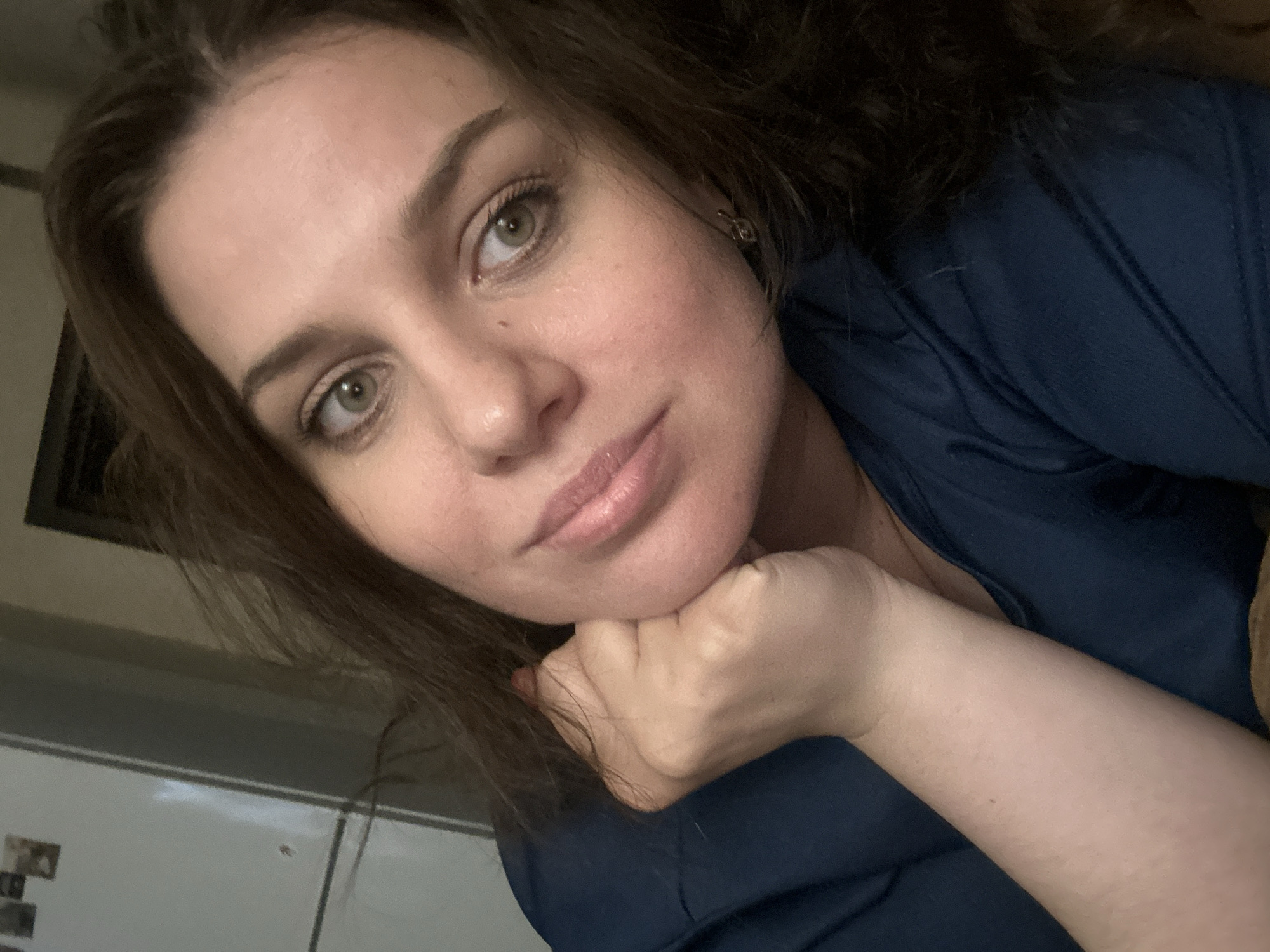A medium shot captures a woman with brown hair, her face turned to the right. She rests her chin on her right hand, with her elbow on her left forearm. The woman wears a dark blue t-shirt and has light-colored skin with rosy cheeks. Her eyes are open and focused to the right of the frame, and her lips are slightly parted. She has light makeup on, including mascara and lipstick. The background is dark and out of focus, with a hint of a white refrigerator on the left. The scene appears to be indoors, in an unknown urban environment.