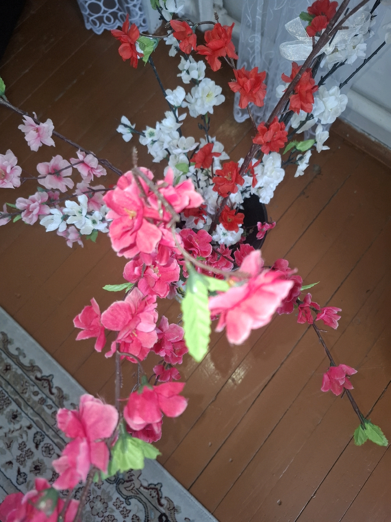 The image depicts a close-up, overhead view of artificial cherry blossom branches arranged in a dark vase. The flowers are a mix of bright pink, white, and red, with some green leaves. The branches are set against a backdrop of a wooden floor and a white lace curtain. A portion of a patterned rug is visible in the lower left corner. The overall scene suggests an indoor setting, possibly a home or a commercial space. There are no people or animals present, and no specific actions or interactions are depicted. The location in Almaty, Kazakhstan, cannot be determined from the visual information provided in the image.