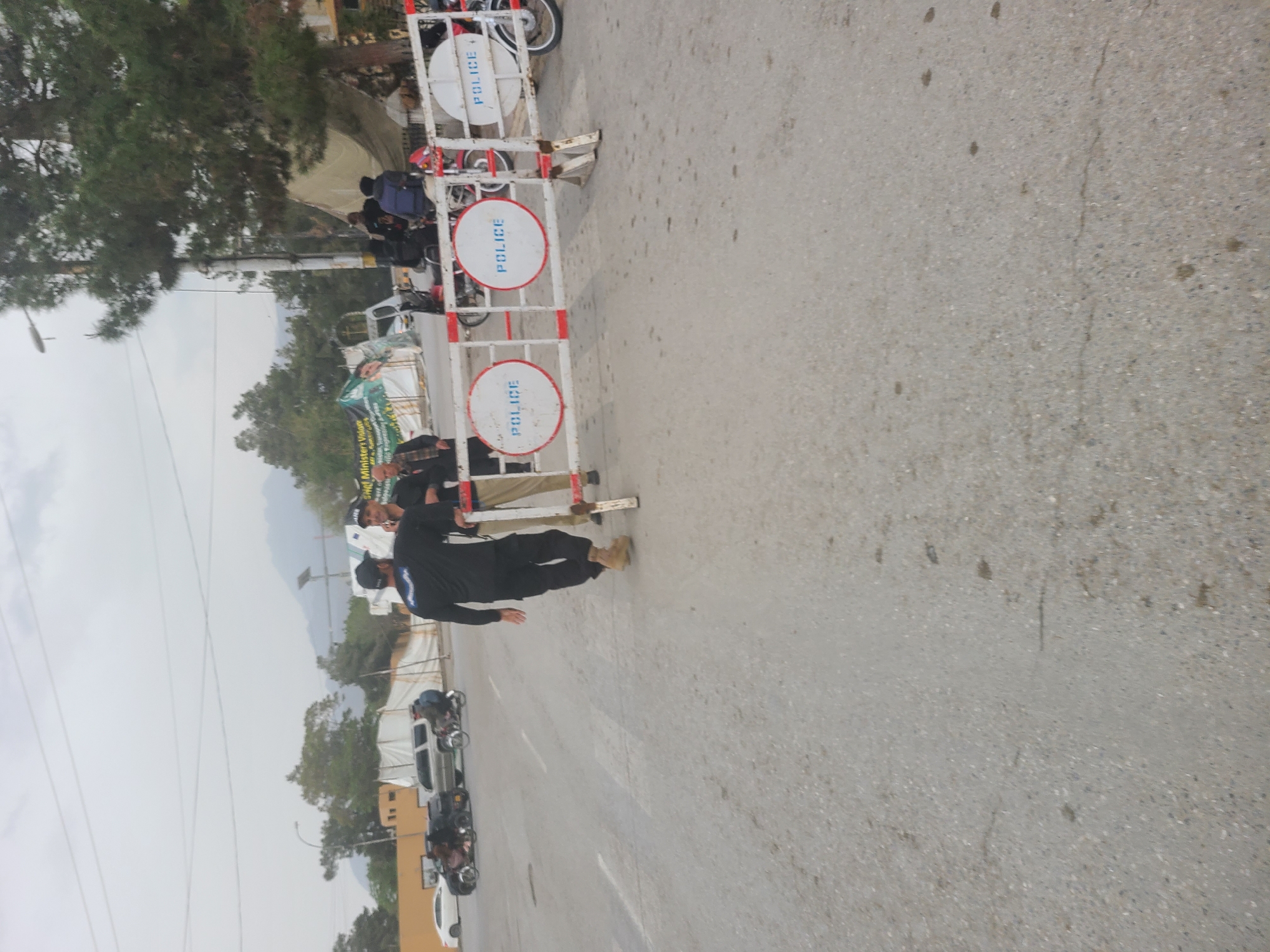 The image depicts a street scene in Quetta City Tehsil, Pakistan. A police barricade with "POLICE" written on it blocks the road. A police officer in a dark uniform steps over the barricade. Other individuals and vehicles are visible in the background, including motorcycles and cars. Trees are present, and the sky appears overcast. The road surface is asphalt.