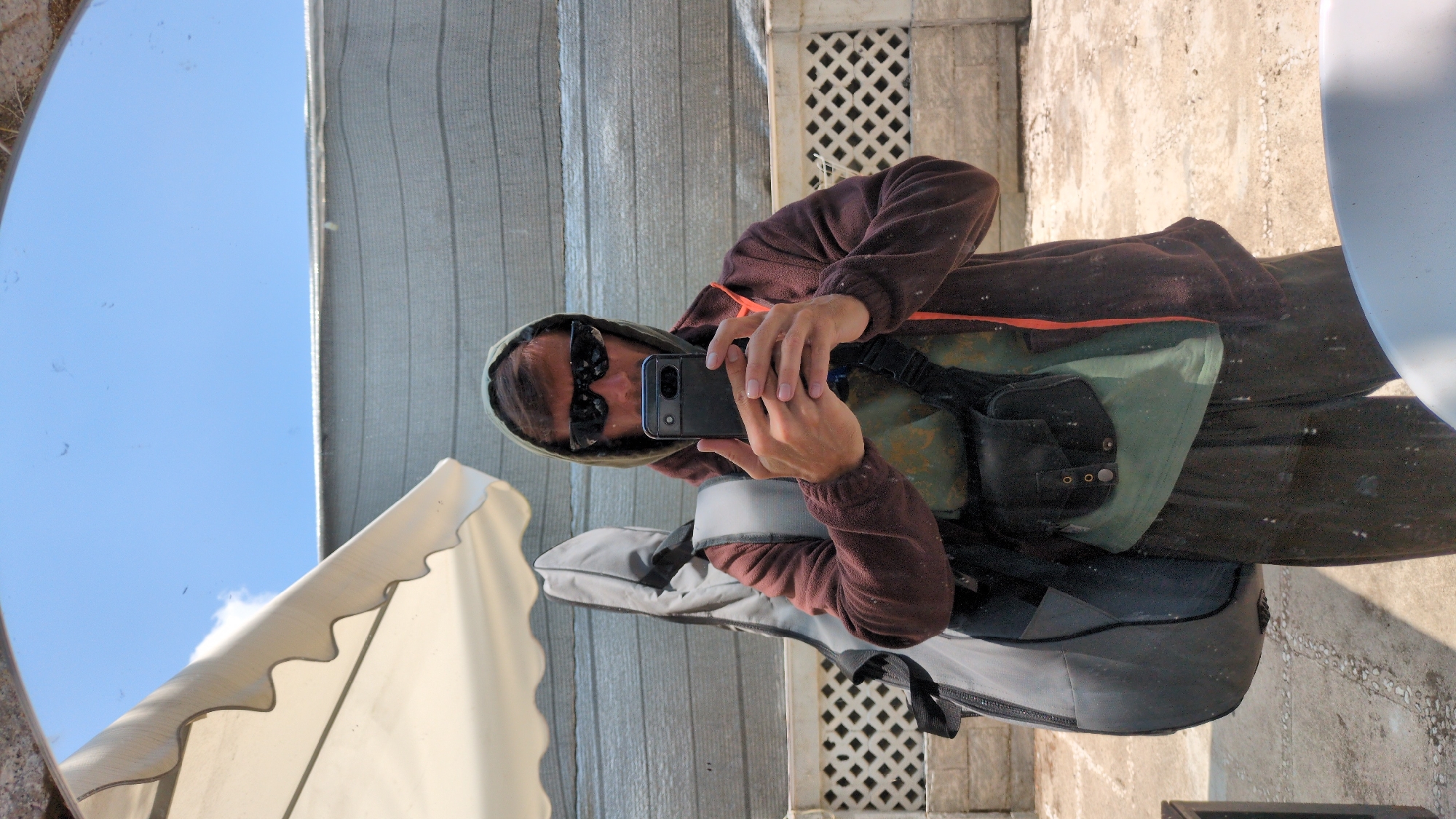 A person wearing a hooded sweatshirt and sunglasses is taking a picture with a smartphone. The person has a crossbody bag and appears to be carrying a guitar case on their back. The reflection is in a round mirror showing the sky and a building. The location is possibly Da Lat, Vietnam, suggested by the background architecture.