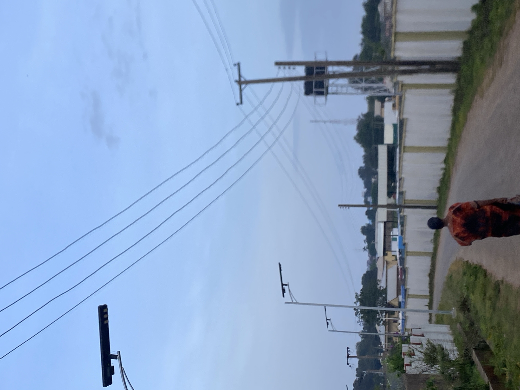 The image depicts a person walking away from the camera on a paved road. The person is wearing a shirt with an orange and dark pattern. The environment appears to be a semi-urban or rural setting, likely in or near Barikin Jirga Summa, Nigeria, given the context. On the left side of the road, there are solar-powered streetlights and some vegetation. A white wall with intermittent green posts lines the right side of the road. In the background, there are several buildings and utility poles with power lines stretching across the sky. The sky is overcast with a soft, diffused light. The scene is calm and appears to be either early morning or late evening.