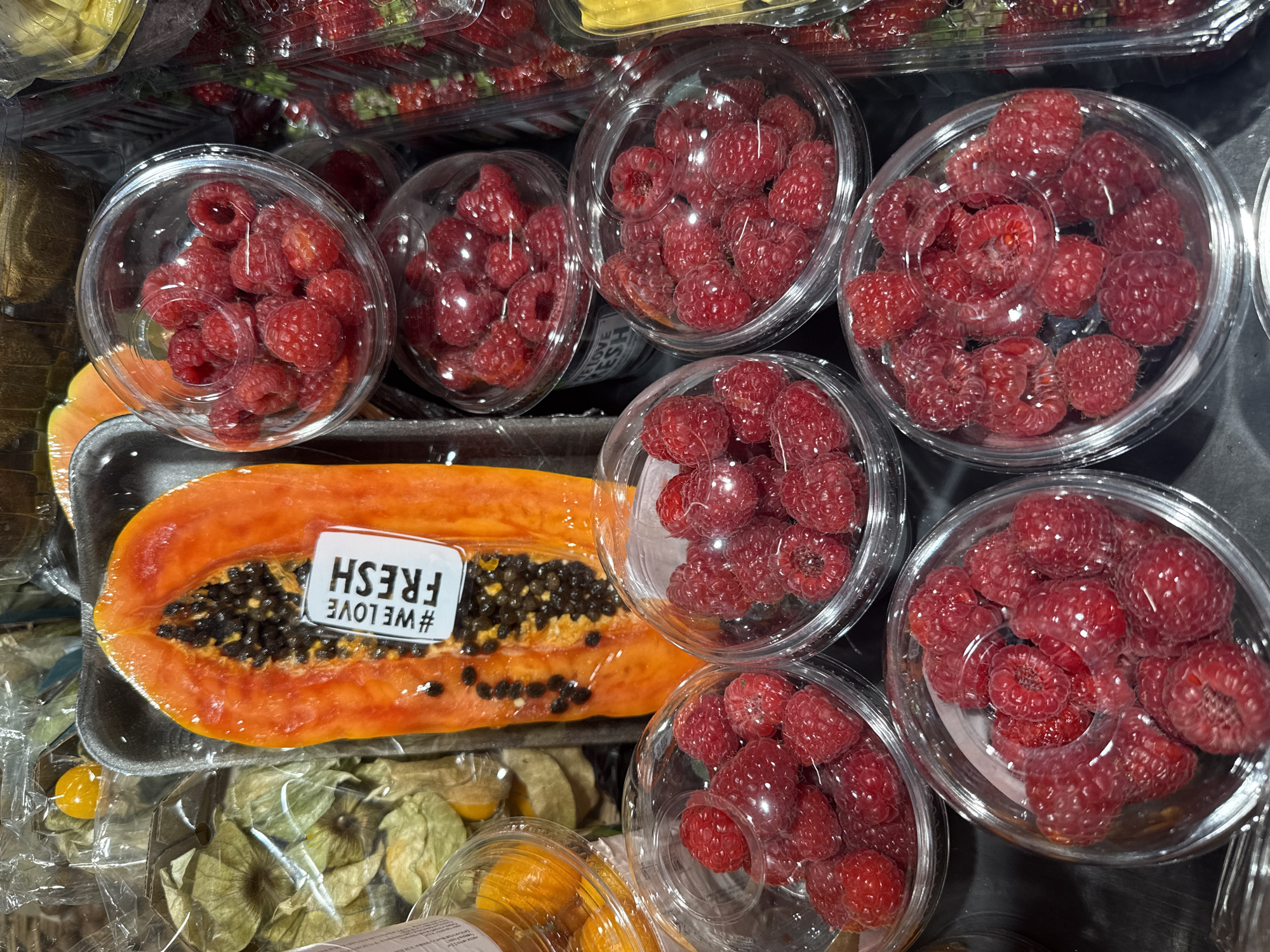 The media file is a close-up, top-down shot of a selection of fresh fruit in a grocery store or market setting. The primary subjects are containers of raspberries and a halved papaya.
There are several clear plastic containers filled with vibrant red raspberries, arranged in a cluster. Each container is filled with a generous amount of berries. To the left of the raspberries is a halved papaya, with its bright orange flesh and numerous small, dark seeds visible. The papaya is on a black foam tray and is partially wrapped in clear plastic. A white sticker with black text is affixed to the papaya, displaying "#WE LOVE FRESH" in a playful, slightly tilted font.
Beneath the papaya and raspberries, other fruits are visible, including what appear to be golden berries or gooseberries in clear plastic packaging, and possibly some yellow fruits like kumquats in a container. The overall impression is one of abundant and fresh produce.
The lighting suggests an indoor setting, likely a well-lit retail environment. There are no people visible in the frame. The focus is entirely on the fruit, highlighting their textures and colors. The image conveys a sense of freshness and appeal, typical of produce displays designed to attract customers. The visible text on the sticker offers a positive marketing message about the quality of the produce.