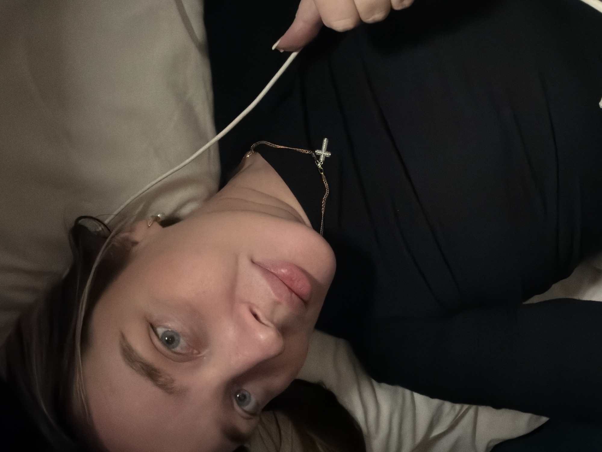 The image shows a young woman lying on her back, with her head tilted to the lower left, looking directly into the camera. She appears to be indoors, possibly in a bed, as she is resting on light-colored, soft bedding or pillows. Her hair is dark brown and cascades around her head.
The woman is wearing a long-sleeved black top. Around her neck, she has a thin gold chain necklace with a small, sparkling silver cross pendant. Her right hand is visible in the upper right portion of the image, holding a white cable, possibly from headphones or a charging device. Her fingernails are painted white.
Her facial features are clear: light blue eyes, well-defined eyebrows, and full lips with a subtle, natural pink hue. Her expression is calm and neutral, with a slight hint of a smile. The lighting in the image is soft, suggesting an indoor environment, possibly in the evening or night. No specific location cues are visible within the image, but the context provided indicates the location is Moscow, Russia. There is no visible text in the image.