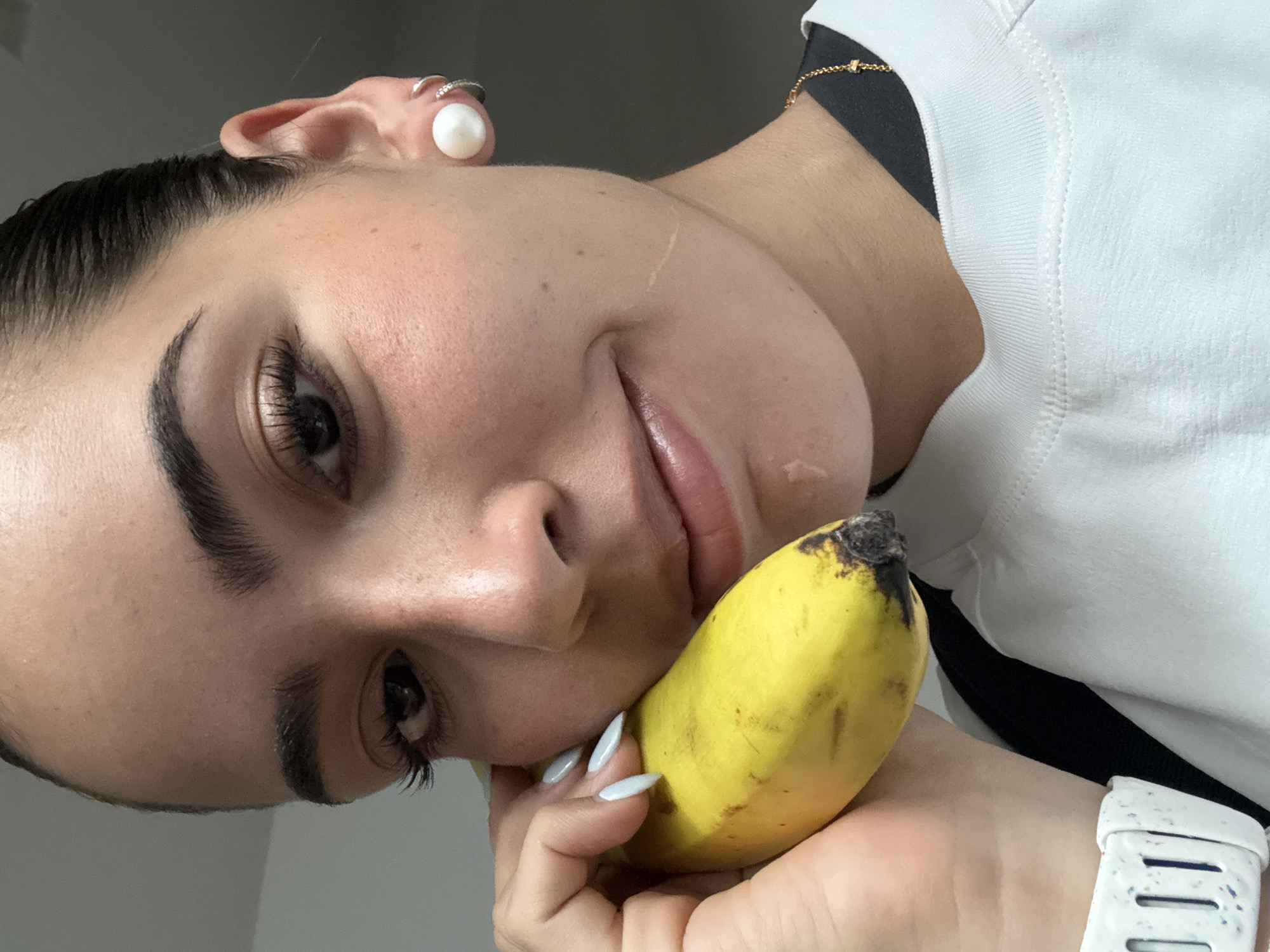 A fair-skinned person with manicured light-blue fingernails is holding a ripe yellow banana, its tip beginning to brown. The person's face is close to the banana. The person has defined eyebrows, long eyelashes, and a pearl earring with other rings on the same ear. A gold chain is visible on the person's neck and a white t-shirt over a black garment is also visible. The person is wearing a white watch. The background is plain and light. The scene's location is not discernible from the image.
