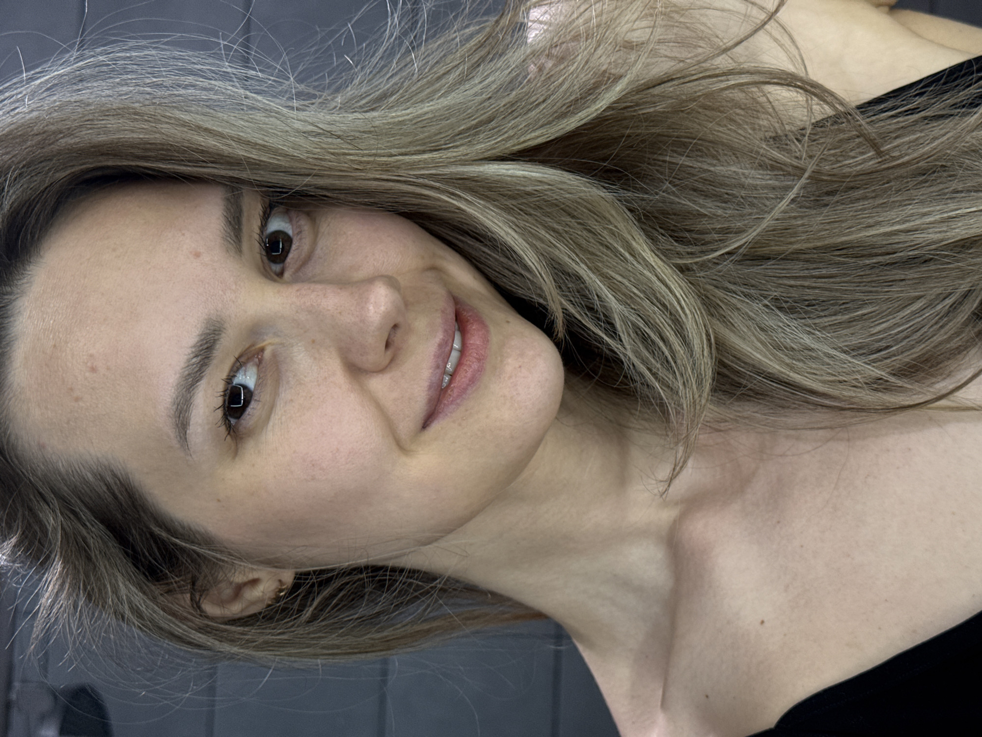 A woman with light brown hair is shown in a close-up shot, angled towards the viewer. She has fair skin and brown eyes, and is smiling slightly. She is wearing a black top. The background appears to be a gray tile wall. There is no indication of the location being Chaikovsky, Russia in the image.