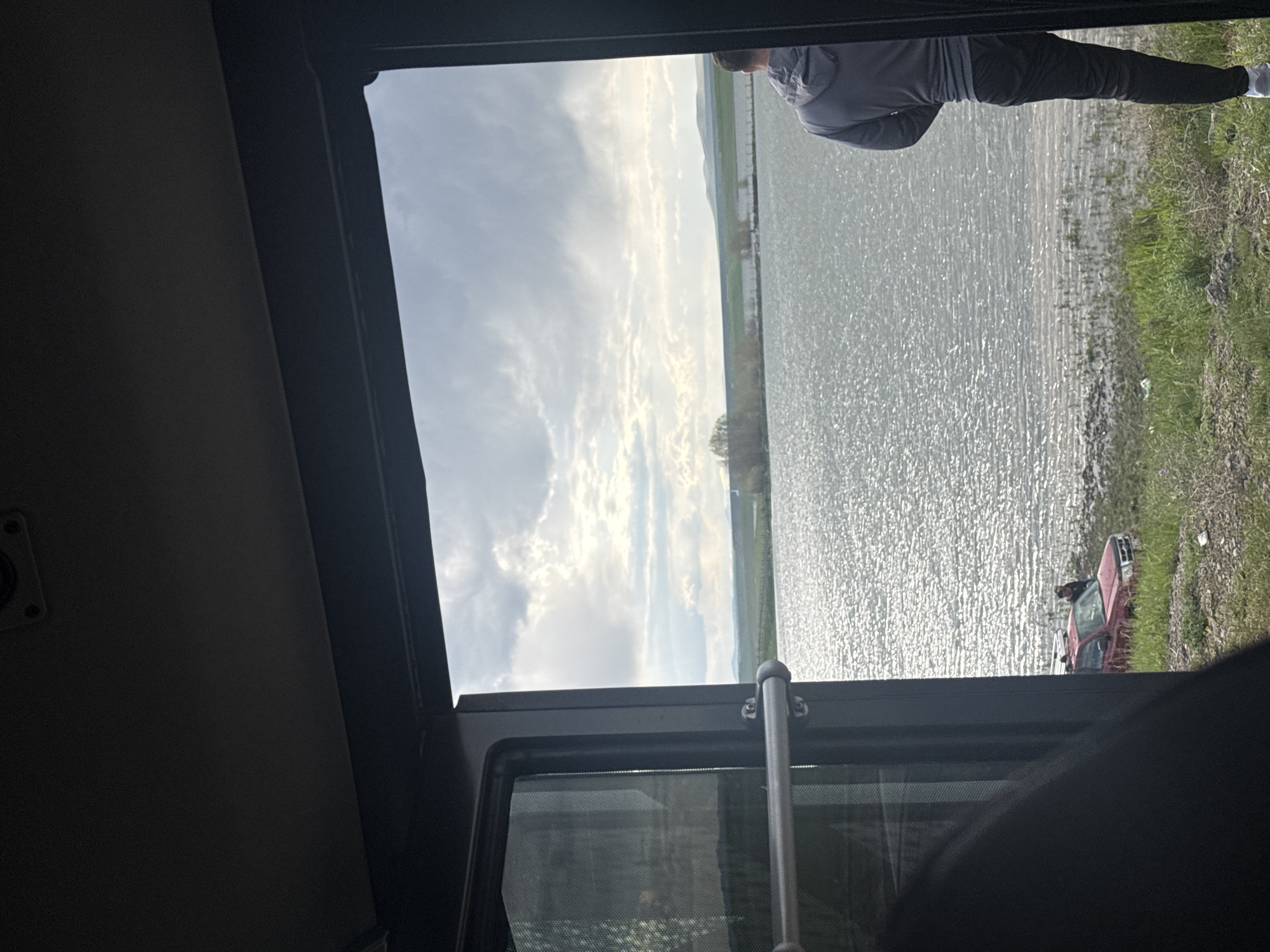 The image shows a view from inside a vehicle, looking out a window onto a body of water. On the shore, a person wearing athletic attire is standing near the water's edge. A red vehicle is parked nearby. The location is not discernible from the image.