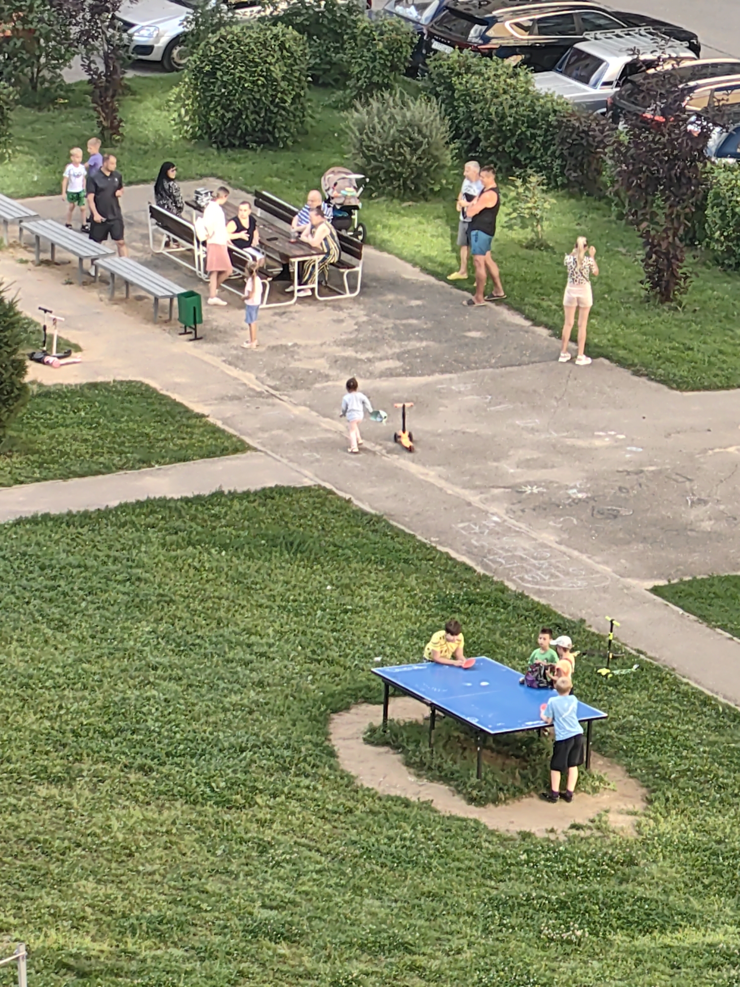 The image depicts an outdoor scene in a residential area, likely a courtyard in the city of Tver, Russia. In the foreground, several children are gathered around a blue ping pong table. One boy in a light blue t-shirt and black shorts is standing, holding a paddle. Three other children are seated or partially visible around the table, one in a yellow shirt and another in a green shirt. A scooter is parked near the table.
Further back, a group of adults and children are seated at picnic tables. Two adults, a man and a woman, are engaged in conversation. A woman in a headscarf is also present. A baby stroller is visible nearby.
In the background, several parked cars are visible, along with lush green bushes and trees. The ground is a mix of paved walkways and grassy areas. Another child is seen walking on the pavement. A woman in a patterned top and shorts is standing alone, facing away from the camera. Two men are standing together on the pavement, one in a black sleeveless shirt and blue shorts, the other in a light blue shirt and shorts.
The overall scene suggests a casual, communal gathering on a pleasant day.