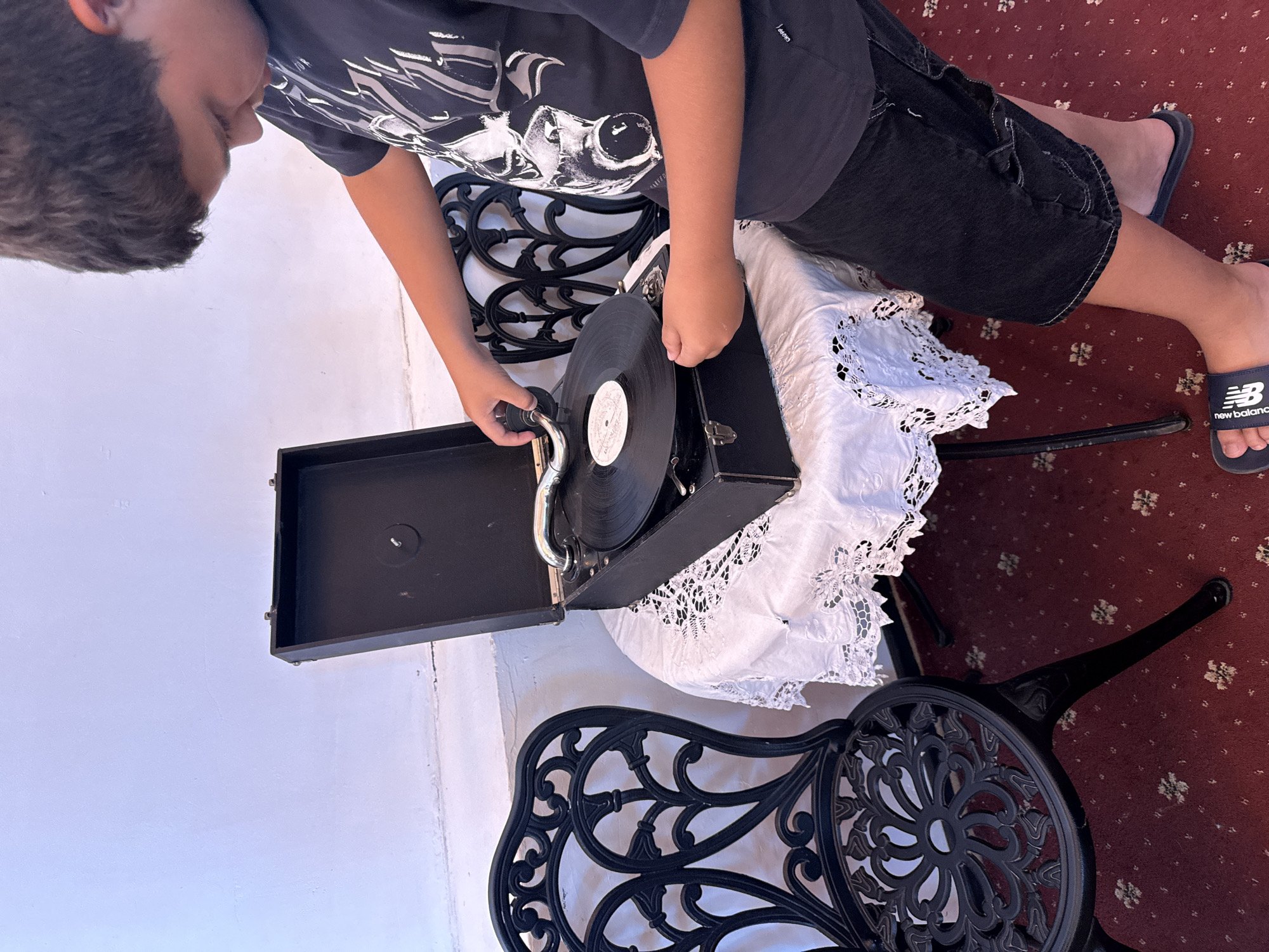 A young person, viewed from above and rotated 90 degrees counter-clockwise, is interacting with an antique record player. They are wearing a dark t-shirt with a graphic print and dark shorts, and are barefoot with one foot wearing a dark sandal with "new balance" written on it. The record player, a portable model with a black case, is open, and a vinyl record is positioned on its turntable. The person's hands are on the record player, suggesting they are either setting up the record or preparing to play it.
The scene is set indoors, indicated by the indoor lighting and the presence of a decorative black metal chair with an ornate design. A white lace tablecloth or runner is draped over a surface near the record player. The background consists of a white wall and a dark red carpet with a floral pattern. The overall impression is one of curiosity and engagement with a vintage object. No specific time of day or weather conditions can be determined. The emotion conveyed is one of focused activity.