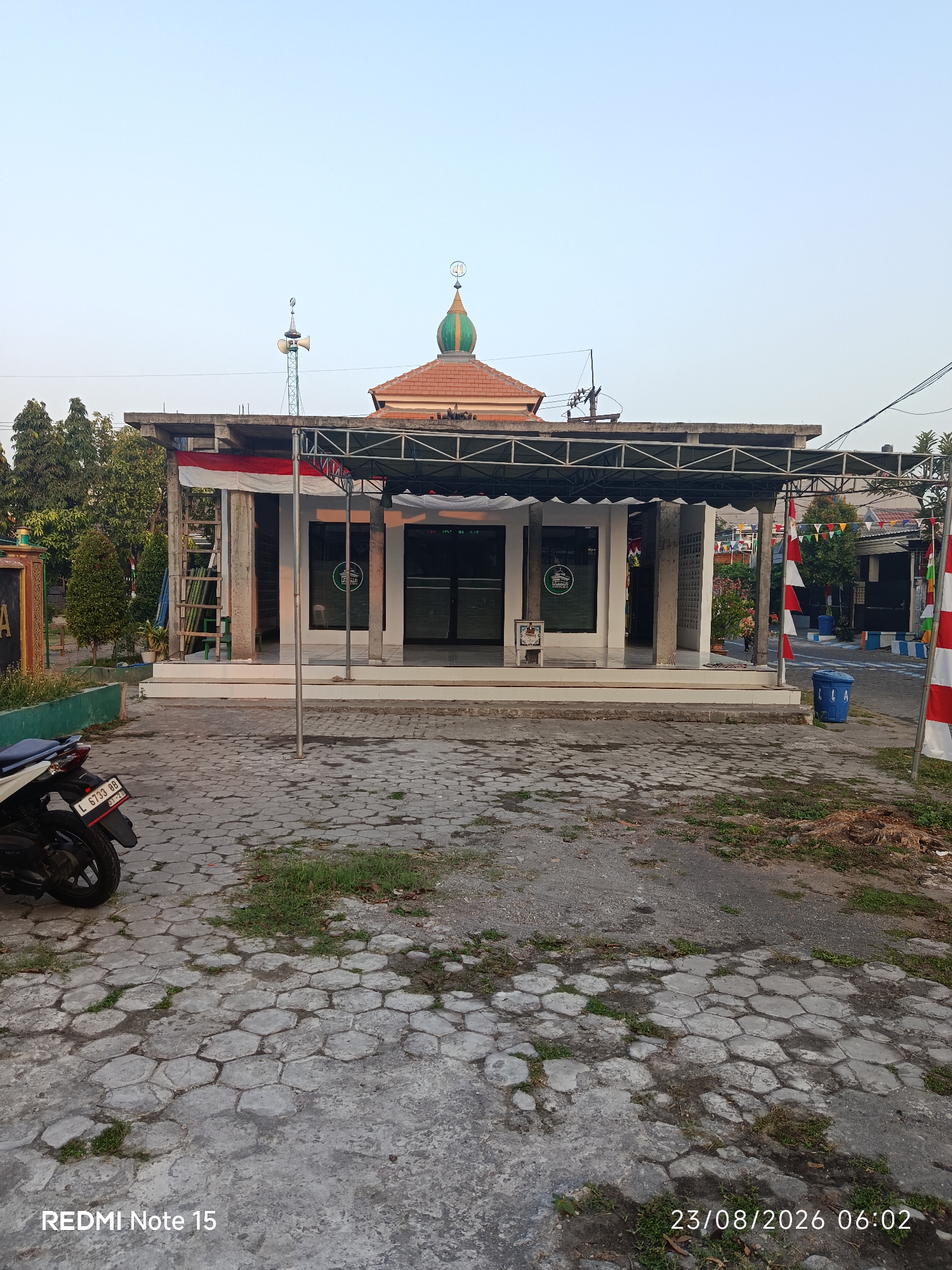 The image shows a building with a dome on top, likely a mosque, located in Keboansikep, Indonesia. The building has a white facade with glass doors and windows. A red and white Indonesian flag is draped over a ladder leaning against the building. Several red and white triangular flags are strung across the street in front of the building. A blue trash can is visible on the right side of the image. The ground in front of the building is paved with hexagonal concrete blocks. A motorcycle is parked on the left side of the image. The sky is clear and pale blue, suggesting it is daytime. The overall scene depicts a religious building and its surroundings in an Indonesian urban or semi-urban setting.