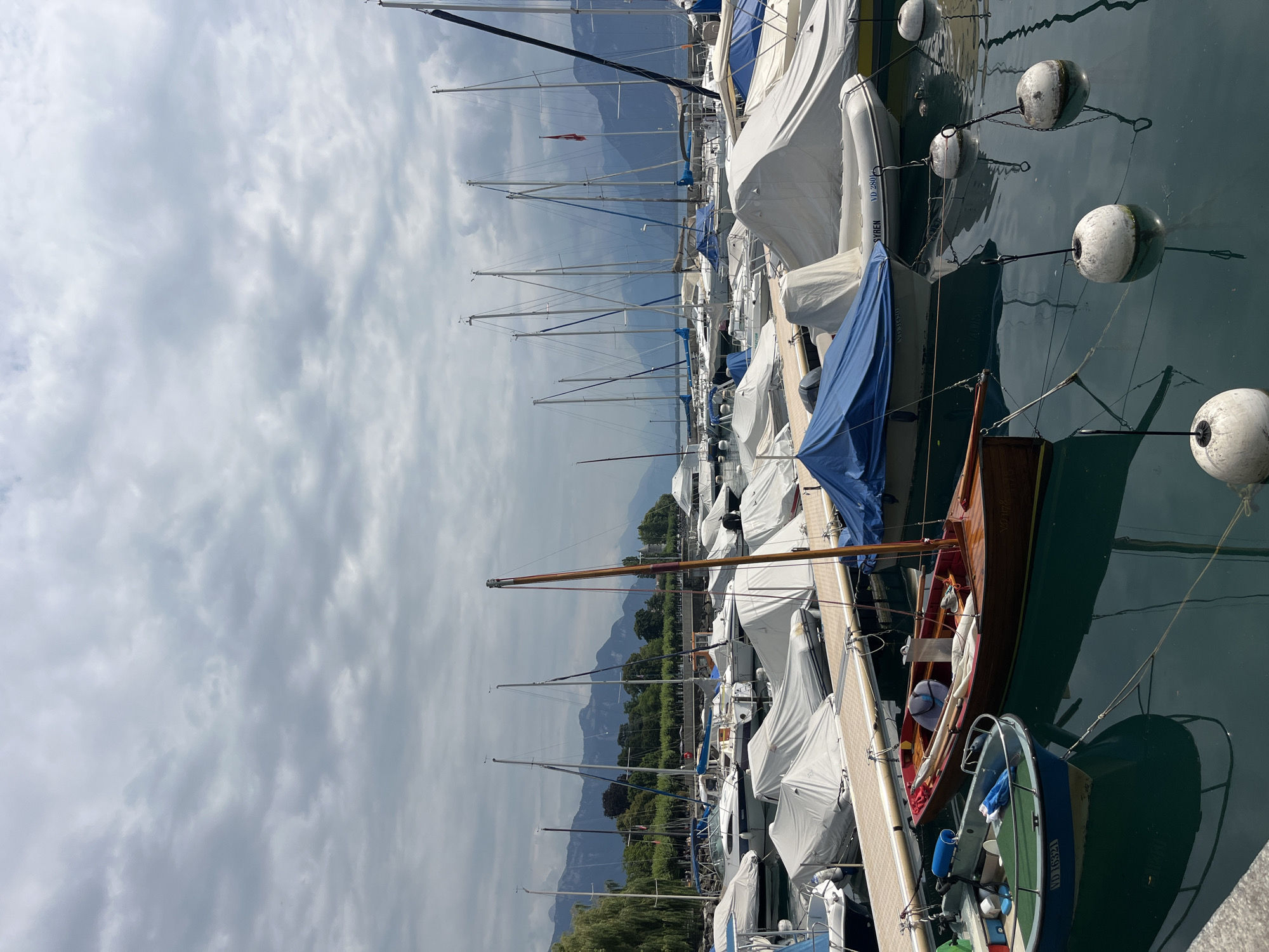 The image shows a harbor in Vevey, Switzerland, with numerous sailboats moored in the water under a cloudy sky. The boats are covered with white tarps, except for a few visible masts and sails. In the foreground, two smaller boats are docked. One is a wooden rowboat, and the other is a blue and green motorboat. The water is calm and reflects the boats and the sky. In the background, mountains and trees are visible. A stone walkway or pier is in the foreground, and there are several buoys in the water. No people are visible in the image.