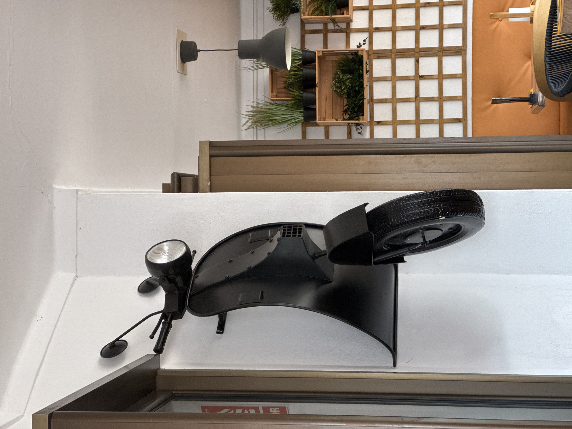 The image captures a scene indoors, depicting a decorative black metal sculpture of a vintage motorcycle affixed to a white wall. The sculpture includes elements like a headlight, mirrors, and a spare tire. The sculpture is mounted above a doorway or window frame with a brown trim.
In the background, on the wall above the sculpture, there's a modern gray adjustable wall lamp. To the right of the lamp, the wall is adorned with wooden crates containing various green plants, adding a touch of nature to the interior. Further to the right, there's an artistic installation featuring a wooden grid against an orange backdrop.
Without additional contextual information, specifying the exact location within the city of Santanyí, Spain, is not possible. However, the interior design elements suggest a modern or artistic establishment.