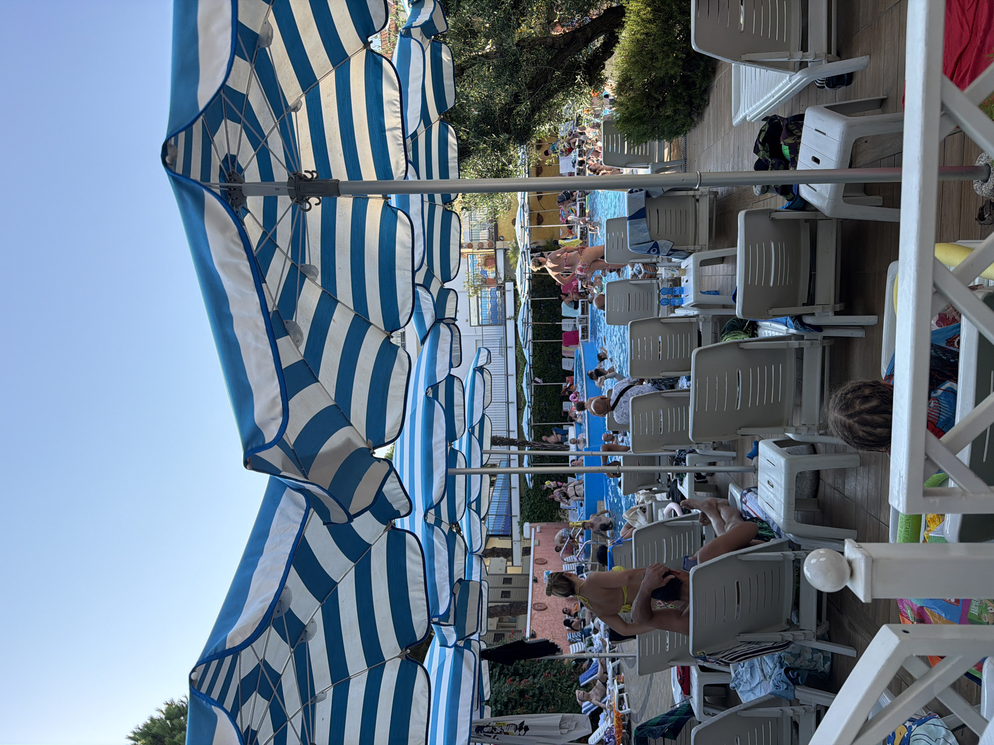 The image depicts a sunny day at an outdoor swimming pool area, likely a resort or hotel, in Gatteo, Italy. The foreground is dominated by the underside of several large blue and white striped umbrellas, casting shadows over rows of empty white plastic lounge chairs. Beyond the chairs, a bright blue swimming pool is filled with people of various ages swimming, wading, and socializing. Some individuals are seen relaxing on the pool deck, either on lounge chairs or standing. The background shows buildings with balconies and potted plants, suggesting a Mediterranean architectural style. Lush green foliage, including trees, borders the pool area. The overall atmosphere is one of leisure and recreation.