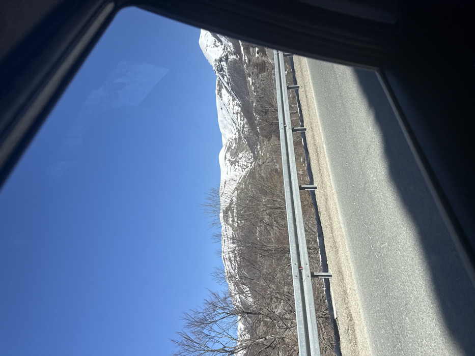 The image depicts an exterior mountain landscape viewed from the passenger side window of a moving vehicle. The scene is located in the vicinity of Godijelji, Montenegro. The foreground shows the dark frame of the vehicle's window and a dark shadow cast by the vehicle's interior across the bottom portion of an asphalt road. Adjacent to the road, a silver metal guardrail with multiple horizontal bars and vertical support posts is prominently featured. Beyond the guardrail, the terrain consists of sparse, leafless deciduous trees and patches of snow on the ground. In the midground and background, large mountains are visible, their upper slopes covered extensively in white snow, contrasting with darker, rocky, and forested lower sections. The sky above is clear and bright blue. There are no people visible, and no discernible actions or interactions are occurring. The environment suggests a cold, clear day.