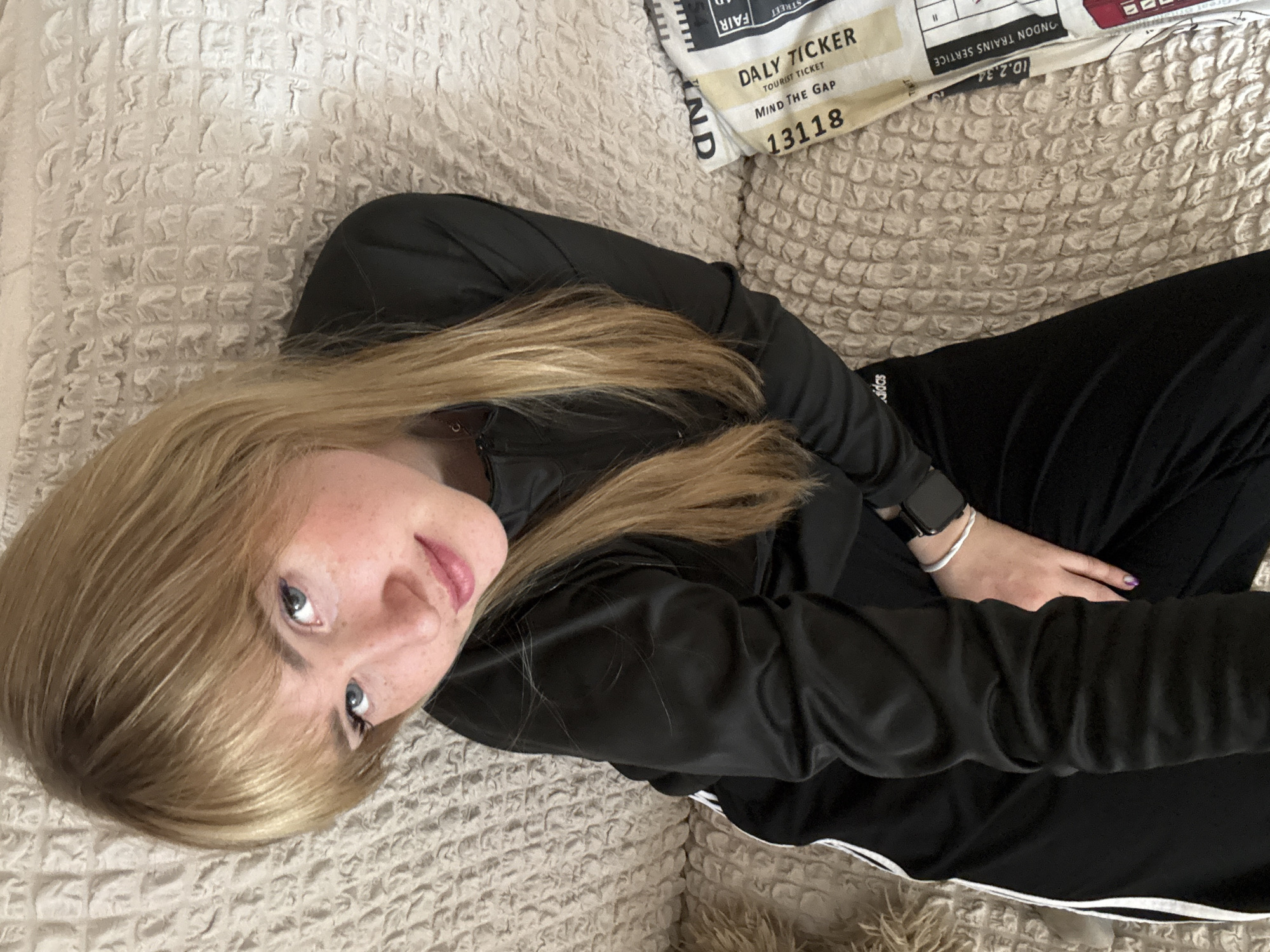 A blonde woman in a black athletic suit and wearing a smartwatch and a bracelet lies diagonally on a beige textured sofa or bed. A pillow with "DALY TICKER" and "MIND THE GAP 13118" printed on it is visible behind her head. There is no location data about Марковский, Russia.