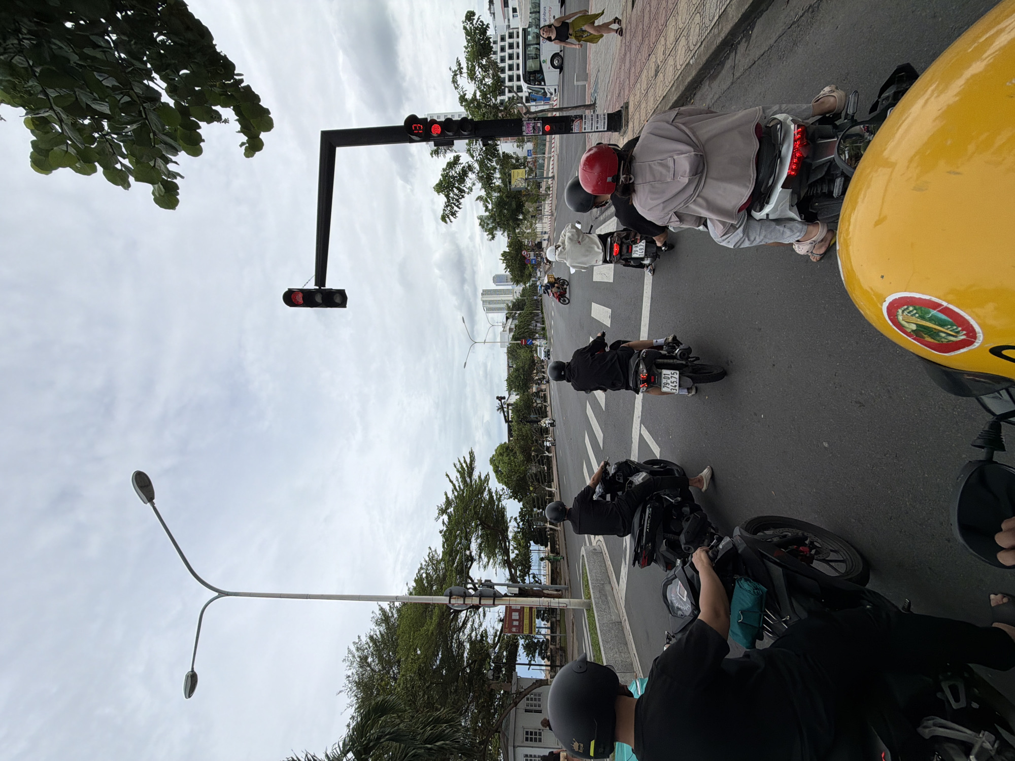 The image shows a street scene in Nha Trang Ward, Vietnam. Several motorbikes and scooters are waiting at a traffic light, which is red. People are riding the motorbikes, wearing helmets. There's a person walking on the sidewalk, and buildings in the background. A street light is visible, and some trees. The sky is overcast.
