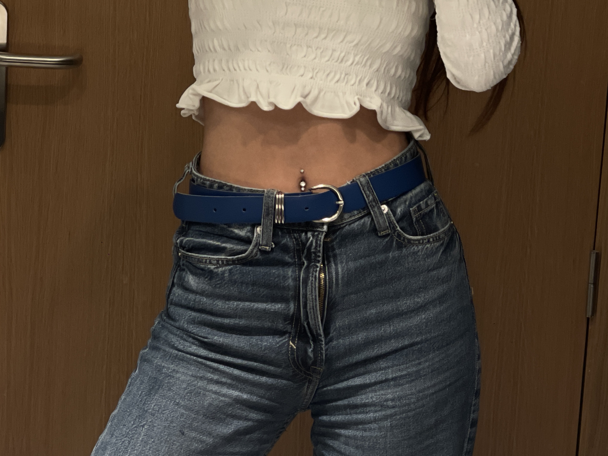 The image shows a person wearing a white, cropped, ruffled top and blue jeans with a blue belt. The jeans have an open zipper. The person has a navel piercing. The background is a plain wooden door.