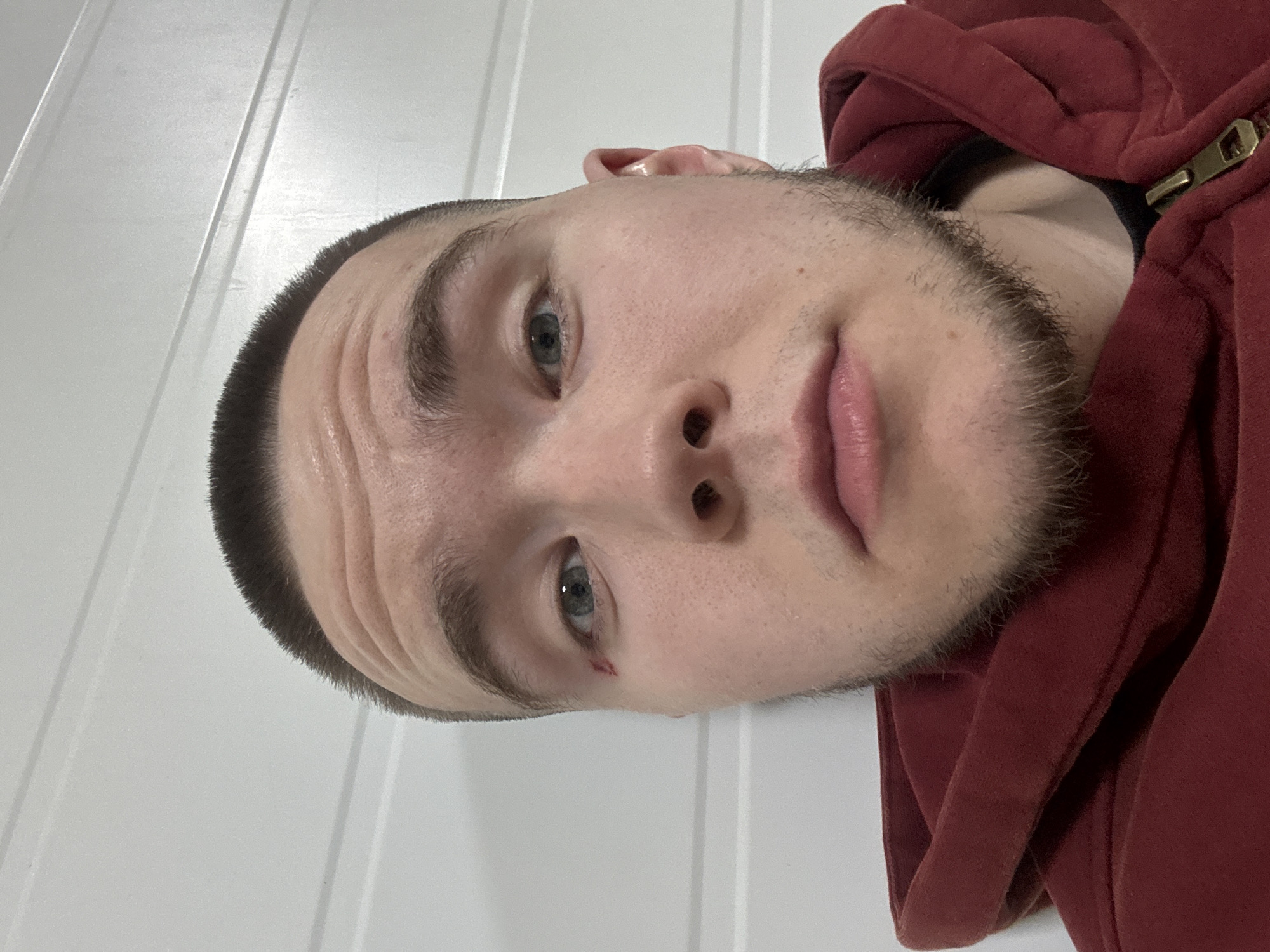 A close-up, angled self-portrait of a young man with short, dark hair and light eyes. He has stubble and a slight cut near his left eye. He's wearing a maroon-colored hooded sweatshirt. The background is a white, paneled wall. The location is not discernible from the image and there is no information to suggest the photo was taken in Kazan, Russia.