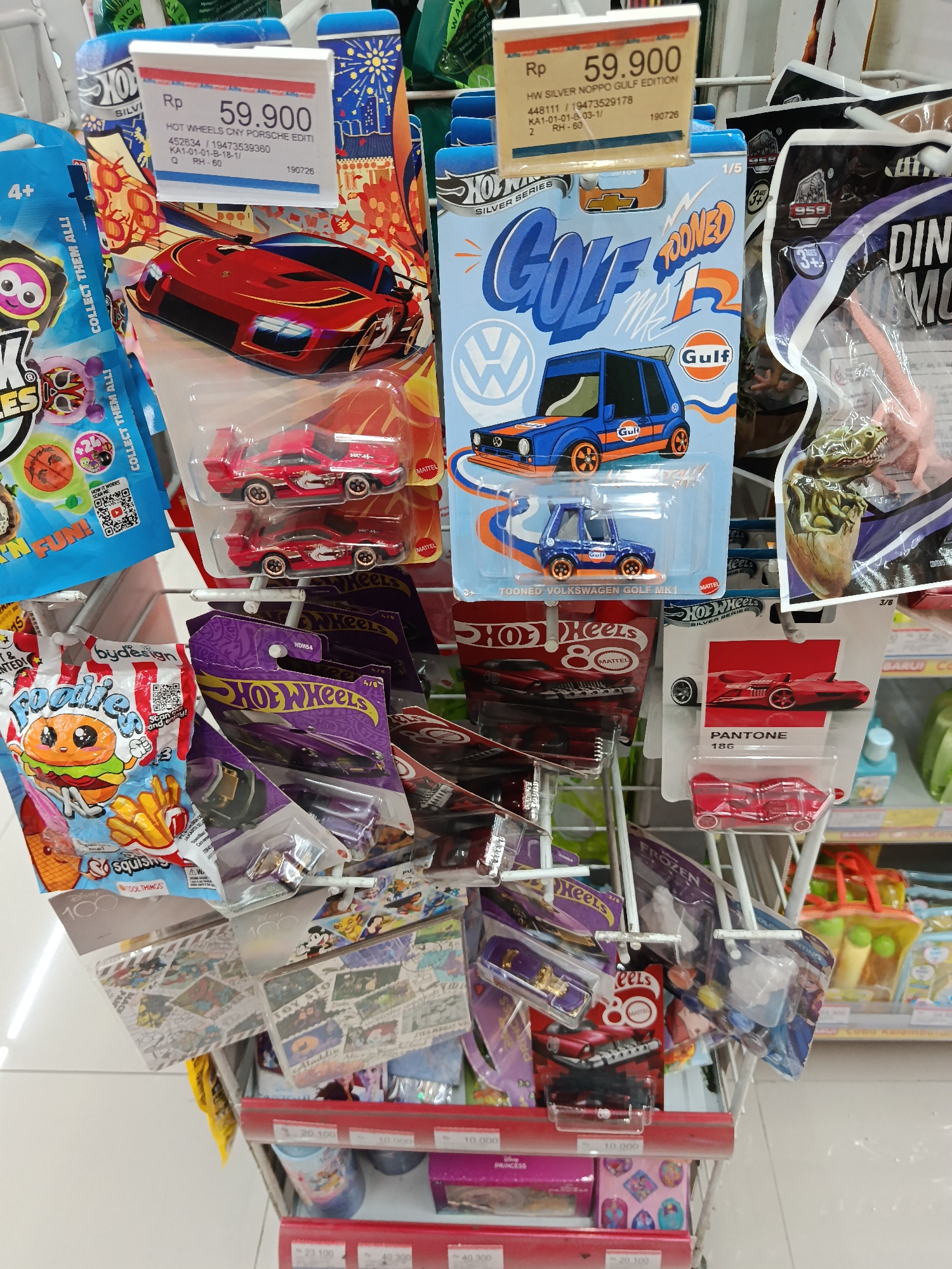 The image is a close-up shot of a retail display shelf filled with toy cars and other packaged toys. The focus is on the Hot Wheels toy cars, some of which are prominently displayed with price tags. The background shows other merchandise, indicating a store environment, likely a toy store or a section of a supermarket. One price tag reads "Rp 59.900" for "HOT WHEELS CNY PORSCHE EDITION". Another Hot Wheels packaging shows a blue Volkswagen Golf with "TOONED GOLF" branding. Further down the shelf, more Hot Wheels cars are visible in their packaging, along with other toy brands. The lighting is bright, typical of an indoor retail setting. The image does not contain any people, animals, or explicit interactions. The scene is consistent with a retail display in Keboansikep, Indonesia, as suggested by the currency used on the price tag (Rp).