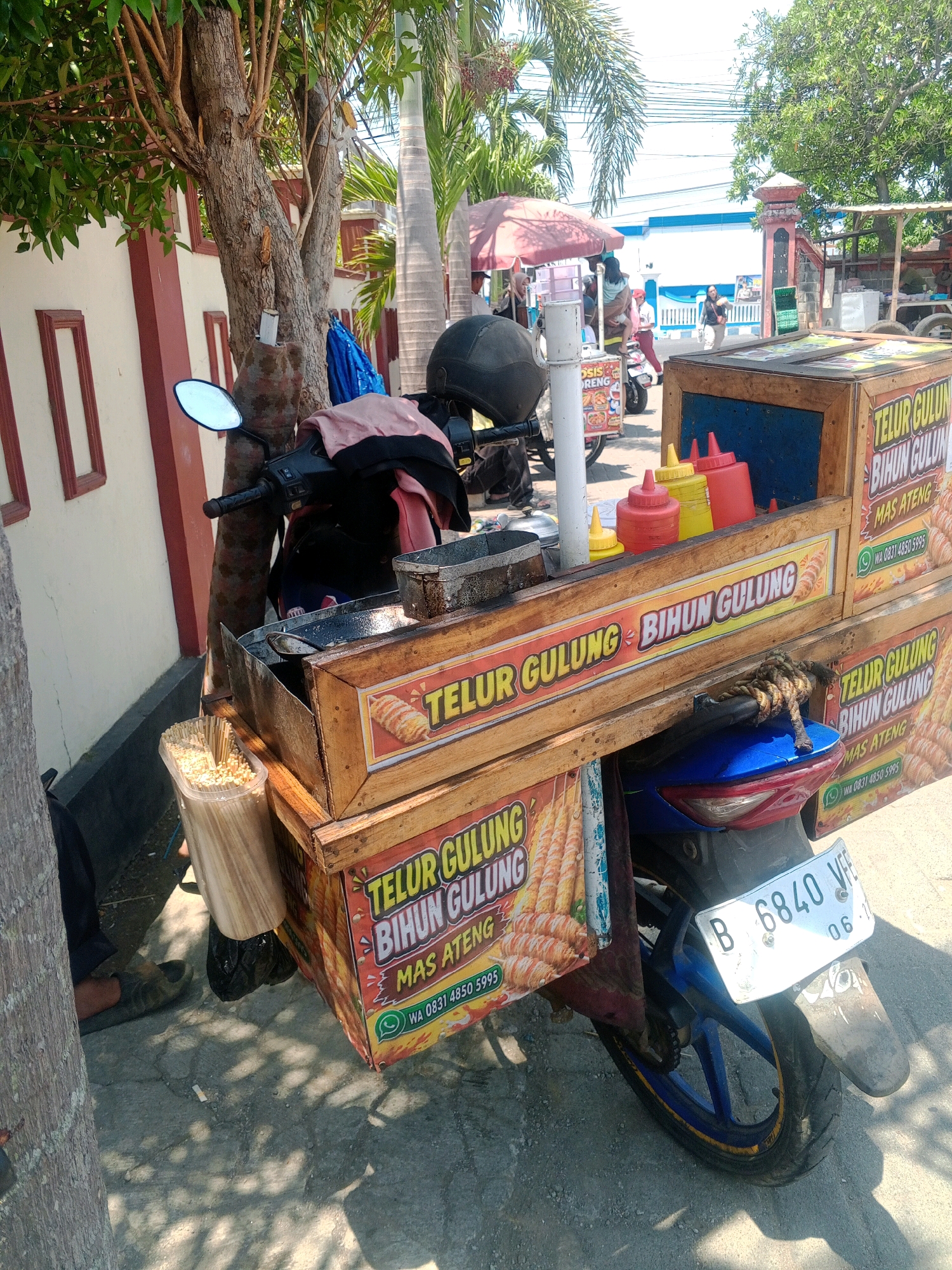A motorcycle street food cart selling "Telur Gulung" (fried rolled eggs) is parked on a street in Giritirto, Indonesia. The cart is attached to a blue motorcycle and is constructed from wood, featuring colorful signage with images of the food item and text in Indonesian. Several bottles of condiments, including red and yellow sauce bottles, are visible on top of the cart. A container filled with wooden skewers for the food is also present. In the background, palm trees and other vegetation line the street. A person's legs and feet are visible on the left side of the image, suggesting they are near or operating the cart. The overall scene depicts a typical roadside food vendor setup.