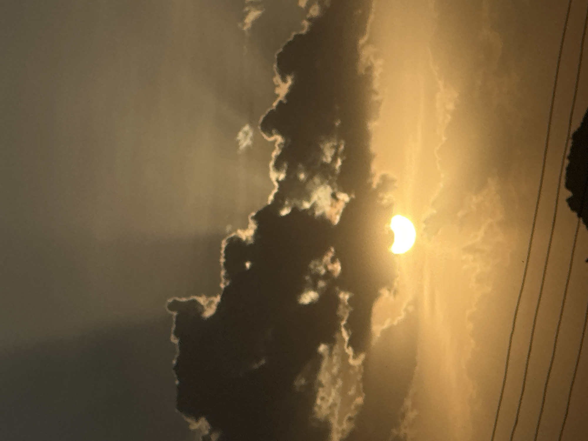The image shows a partial solar eclipse viewed from Bukuru, Nigeria. The sun is partially obscured by clouds. Power lines are visible in the lower right of the frame.