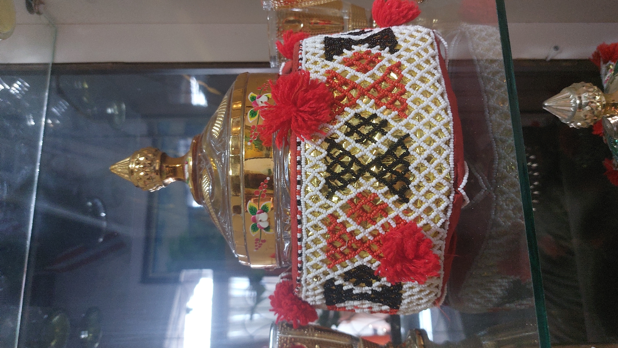 A decorative, lidded container with a beaded, patterned base and a gold finial sits on a glass shelf in what appears to be a display cabinet. The base is adorned with a lattice pattern of white, black, and red beads against a gold background, with red pom-pom-like embellishments protruding from the top edge. The lid is made of clear glass with gold accents and a decorative gold knob. The background is blurred and shows reflections, suggesting an indoor environment. The scene is devoid of people. This object is likely a decorative piece or a container for small items. There is no information to suggest this image is located in the City of Medan, Indonesia.