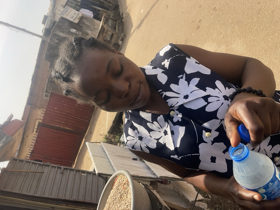 An outdoor, brightly lit, slightly high-angle shot depicts an individual in an urban setting, consistent with the street environment of Jos, Nigeria. The specific street or landmark within Jos is not identifiable.
The primary subject is an adult female, positioned centrally, with her head tilted downwards and eyes closed or looking down, focusing on an object in her hands. She has dark skin and her dark hair is styled in multiple small braids or twists. She wears a sleeveless top with a dark blue or black background and prominent white floral patterns, fastened with light-colored buttons down the front. A dark, beaded bracelet is visible on her right wrist.
In her hands, she holds a transparent plastic bottle, likely containing water, with a blue screw-top cap. Her right hand grips the cap, suggesting an action of opening or closing the bottle, while her left hand supports the lower part of the bottle.
To the left of the subject, partially visible, is a grey, shallow bowl filled with an assortment of grains, appearing to be a mix of light-colored beans and smaller, possibly red, grains. This bowl rests on an elevated surface, possibly a market stall or vendor's stand, next to what appears to be white fabric or dried goods hanging vertically.
The background shows a dusty or unpaved road surface. Further back, a tall, reddish-brown corrugated metal gate with vertical bars is visible, leading into what appears to be a residential or commercial property. Beyond the gate, multiple weathered, light-colored buildings with signs of disrepair are present under a clear sky. A wooden structure, possibly another stall or a bench, is discernible in the far right background, with a white garment hanging on it. The scene is illuminated by strong sunlight, casting minimal shadows.
