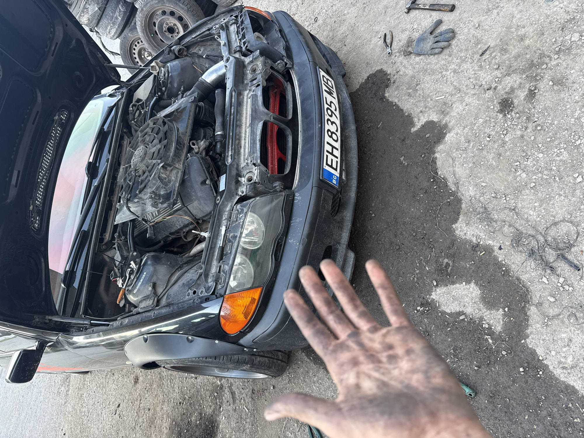 The image depicts a black car with its hood open, revealing the engine. The front of the car faces the right of the frame and is angled slightly towards the viewer. The Bulgarian license plate "EH 8395 MB" is visible. A dirty hand is extended towards the car. The environment appears to be an outdoor work area, possibly a mechanic's yard. Tools such as a hammer and pliers, a glove, spare tires, and other car parts are scattered around. The location is likely Shumen, Bulgaria, based on the license plate and the user prompt.