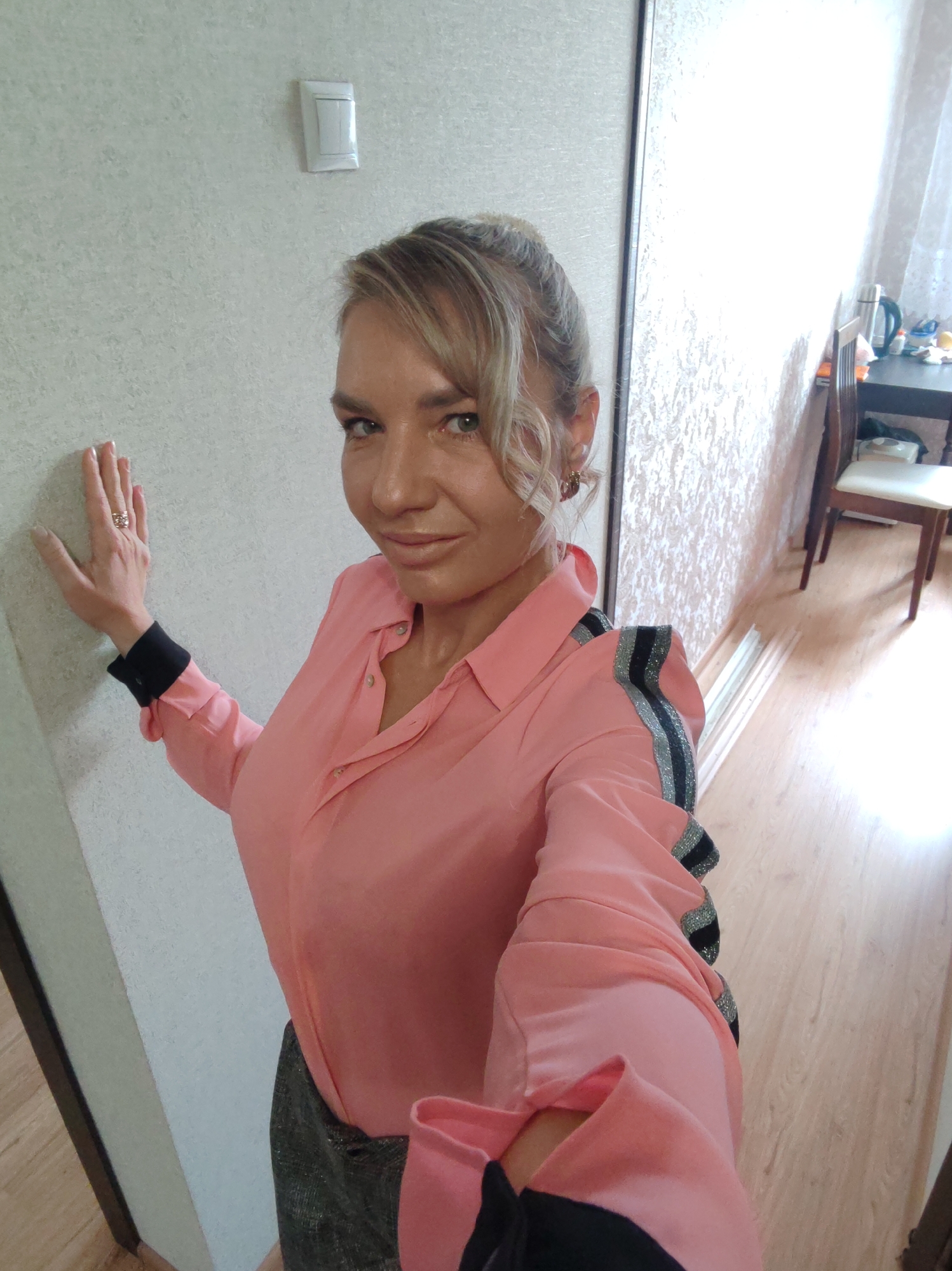 This media file is a self-portrait of a woman standing indoors, leaning against a light-colored, textured wall. She is wearing a pink collared shirt with black and silver striped accents on the shoulders and black cuffs. A sparkly dark skirt is visible below the shirt. Her right hand is pressed against the wall, and she is wearing a ring on her index finger. Her hair is blonde and styled with some curls around her face. Her expression appears neutral to slightly pleased.
The background shows a hallway with a wooden floor. To the right, there is a doorway leading to another room. Inside that room, a table is set up with a kettle and some other items. A chair with a white cushion is also visible. The lighting suggests it could be daytime, as light streams in from the right. The overall setting appears to be a residential interior. There is no visible text in the image. The location context is Petropavlovsk-Kamchatsky, Russia.