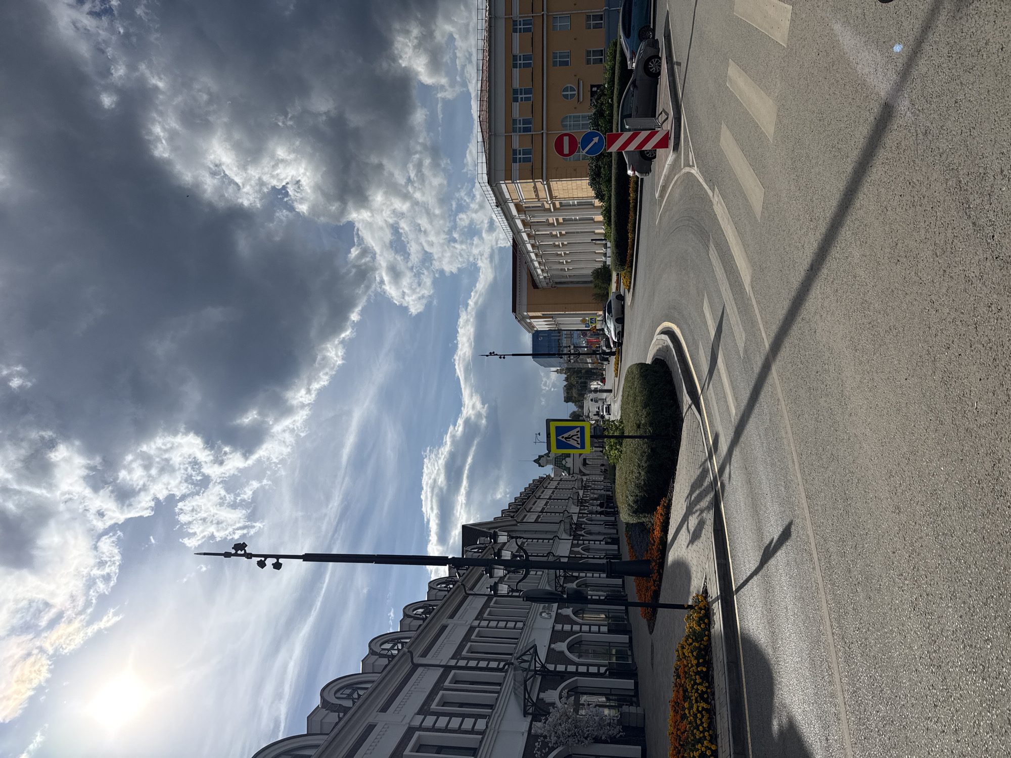 The image captures a street scene in Perm, Russia, on a day with dramatic, cloudy skies and bright sunlight breaking through. On the left, a long, multi-story building with a white facade and dark arched windows lines the street. Decorative dark trim accentuates the windows and roofline. Planters filled with orange and yellow flowers are visible along the sidewalk. A tall, black lamppost with multiple lights stands prominently in front of the building.
The street curves into the distance, revealing a larger, yellow building with a row of white columns and numerous windows on the right side of the frame. A pedestrian crossing is marked on the asphalt road, with a yellow pedestrian crossing sign nearby. Traffic signs, including a "no entry" sign and a blue directional arrow sign, are positioned on a red and white striped post at the intersection. Several cars are parked along the street and driving on the road. The overall impression is a well-maintained urban environment with a mix of architectural styles under a dynamic sky.