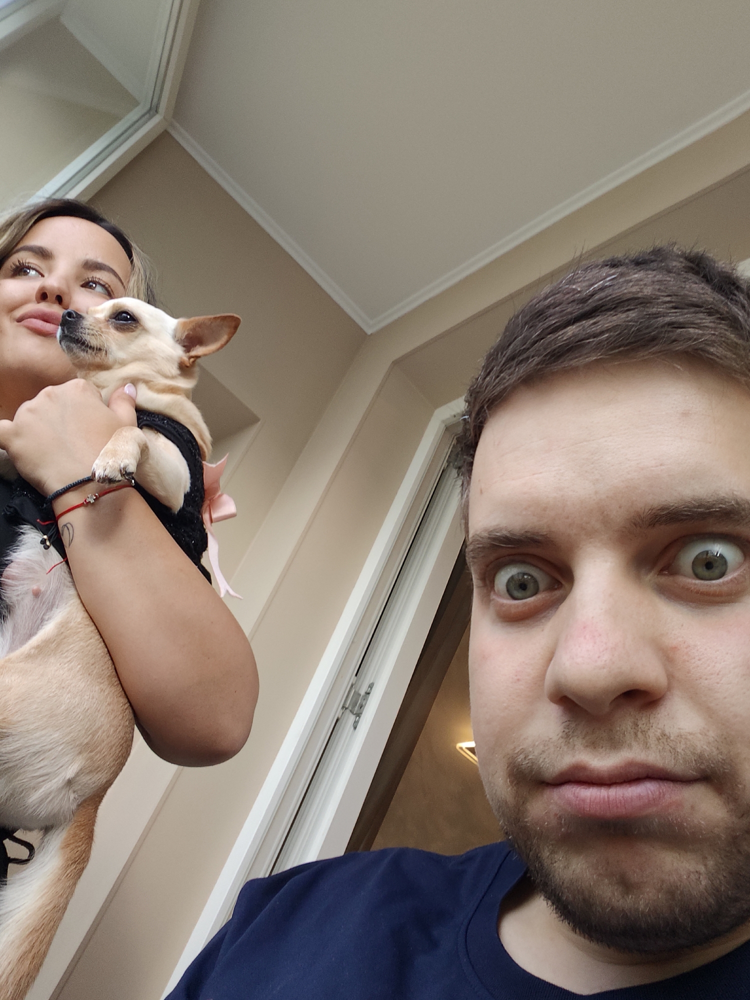 This casual, low-angle shot captures an intimate indoor moment featuring two people and a small dog, set in what is described as Sofia, Bulgaria. The scene appears to be well-lit by natural light, suggesting it is daytime.
In the foreground, a man with short brown hair and light facial stubble looks directly at the camera with a wide-eyed, almost startled or playful expression, his lips pursed. He is wearing a plain dark blue crew-neck t-shirt.
To the left and slightly behind the man, a woman is partially visible from the chest up, holding a small, light-colored dog. She has long, light brown hair and is looking upwards and to the left, away from the camera, with a soft, content smile. On her left wrist, she wears a red string bracelet with a small charm and a black string bracelet, along with a small heart tattoo on her forearm.
The dog, a small breed like a Chihuahua with fawn-colored fur, is cradled in the woman's arms and is also looking in the same direction as the woman, away from the camera. It is wearing a black garment or harness adorned with a delicate light pink bow.
The background shows light-colored walls and a ceiling with white molding. A window or glass door frame is partially visible, confirming the indoor setting and the presence of natural illumination. The overall mood of the image is candid and warm. There is no visible text in the media file.
