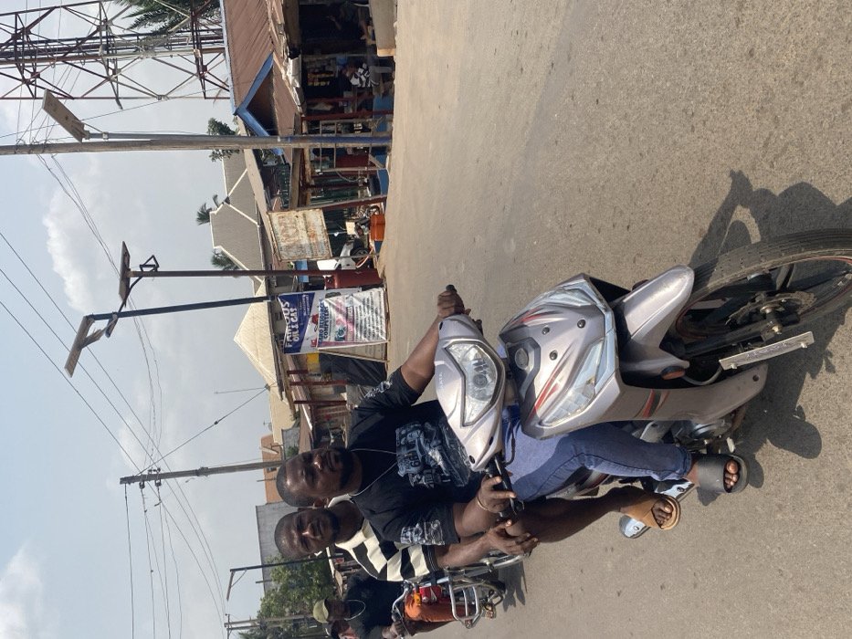 A daytime street scene in Afikpo, Nigeria, captures two adult males on a light gray scooter-style motorcycle, in motion on a paved road. The primary subject, driving, is a male wearing a dark t-shirt with a graphic design, blue jeans, and black slides. He holds the handlebars. A male passenger, wearing a horizontal striped t-shirt and light-colored sandals, rides directly behind the driver. Behind them, a third male operates a red motorcycle.
The environment includes overhead utility lines and multiple dark-colored poles supporting solar-powered streetlights. To the right, a series of single-story commercial buildings with corrugated metal roofs line the road. Prominent among these is a business identified by a large sign displaying "FAMOUS OIL & GAS" in blue text on a white background, positioned next to a red fuel dispenser. Another sign, "TARO RESOURCES COMPUTERS," is visible nearby. Orange jerry cans are situated on the ground near the "FAMOUS OIL & GAS" establishment. In the far background to the right, a tall metal lattice tower (likely a telecommunications mast) stands against a bright, hazy sky. Other individuals are present near the commercial structures. Strong shadows are cast on the road surface, indicating direct sunlight.