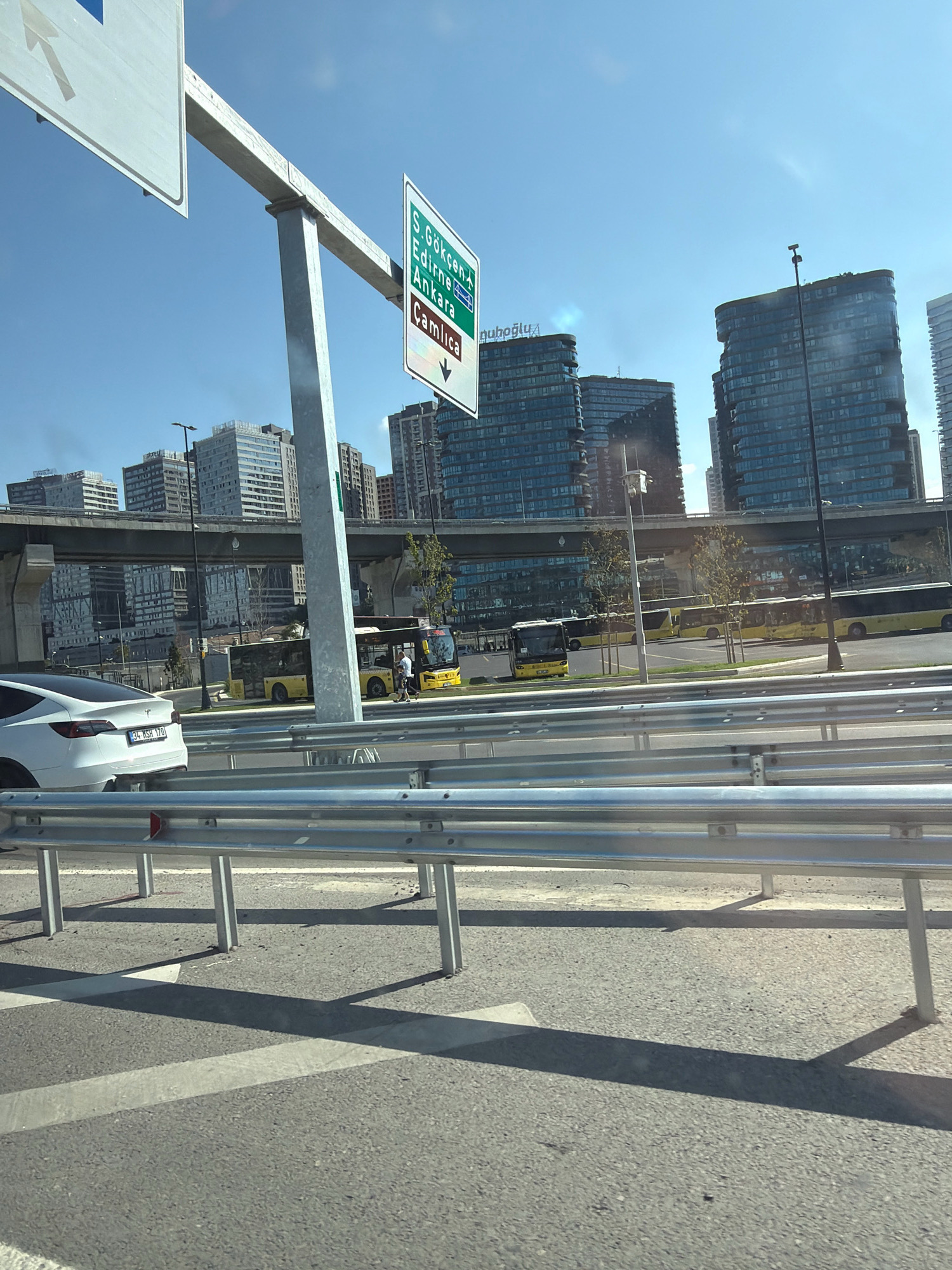 A white Tesla Model 3 is parked on the right side of the road. The license plate is visible and reads "34 RSR 170". In the background, several yellow buses are parked, and a man is walking between two of them. The scene is set in Kadıköy, Turkey, with modern skyscrapers visible in the distance. A highway sign indicating "S. Gökçen, Edirne, Ankara, Çamlıca" and "Nuhoglu" is also present. The sky is clear and blue, and the sun is shining, casting shadows on the road.