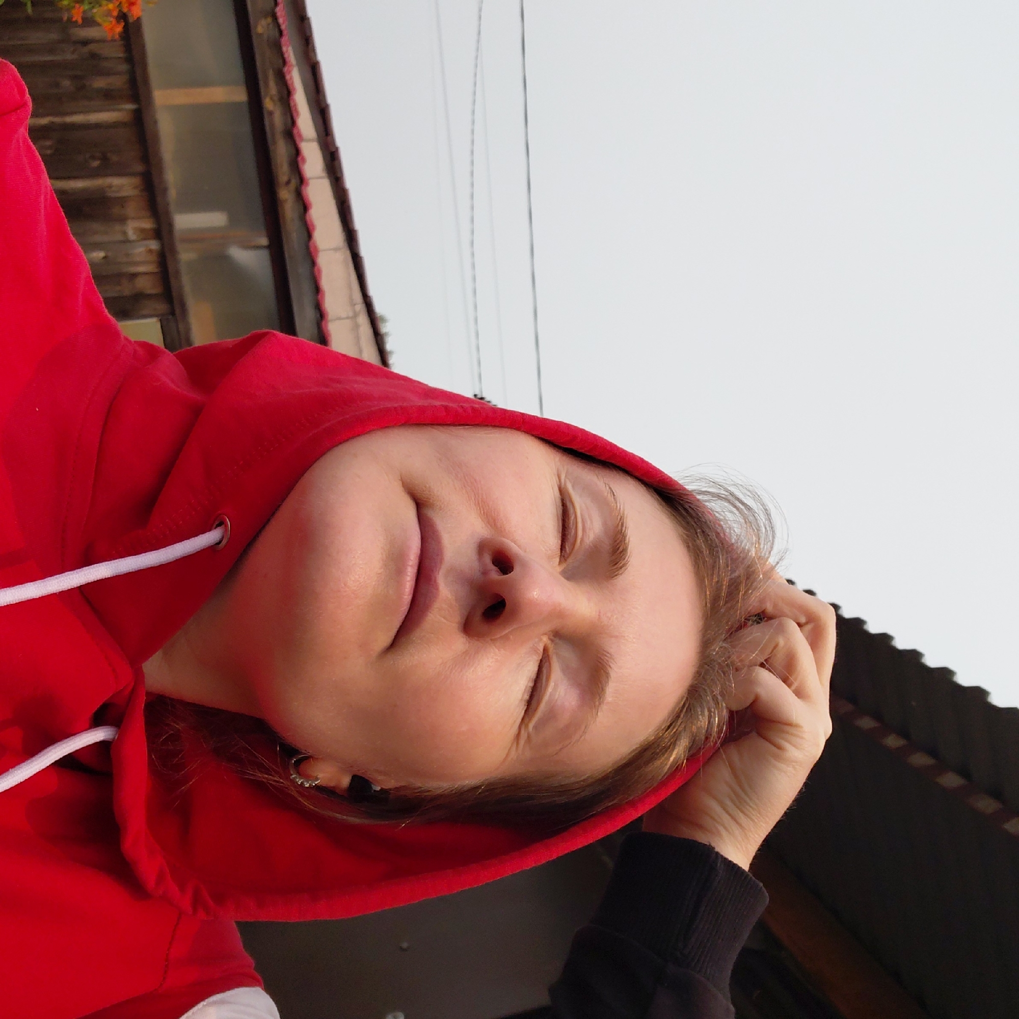 A medium shot photograph shows a woman with her eyes closed, wearing a red hooded sweatshirt. Her right hand is in the hood, scratching the top of her head. Her left hand is not visible. She is wearing a small hoop earring in her visible left ear. In the background, a wooden fence and the roof of a building are visible. The sky is overcast. The location is unknown.