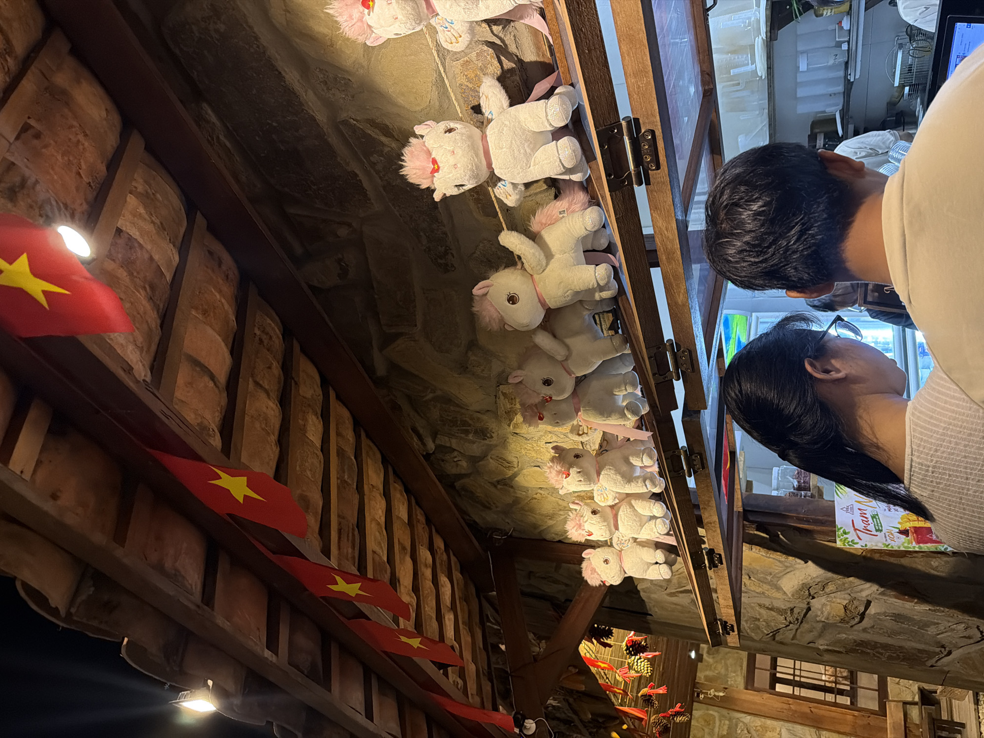 The scene depicts an indoor area, confirmed to be in Vietnam by the presence of multiple national flags (red field with a yellow five-pointed star) strung across a wooden-beamed ceiling. The exact location within Nam Nha Trang Ward is not determinable from the image.
In the foreground, two individuals are positioned with their backs to the viewer, facing a counter. The person on the left is female, with dark hair tied back, wearing glasses and a light-colored, textured top. The person on the right is male, with short dark hair, wearing a light-colored t-shirt. Both individuals are looking downwards, presumably at the counter or an activity behind it.
Above the counter, a wooden shelf displays a line of approximately nine white plush unicorn toys, each featuring a pink mane, tail, and ribbons, with some possessing small wings. The shelf is mounted against a rustic stone wall composed of irregular brown and grey stones.
The ceiling above is constructed with dark wooden beams and terracotta-colored tiles or panels. Multiple light fixtures illuminate the space. On the left side of the image, decorative clusters of pine cones, some adorned with red ribbons, are suspended from the ceiling.
Behind the counter, partially obscured by the foreground subjects, clear plastic containers and pitchers are visible on shelves, indicating a food or beverage preparation area. A person in a white top can be seen working behind the counter. A screen, possibly a point-of-sale system, is visible on the counter to the right. A partially visible sign with Vietnamese text, including "Trâm" and "Xoài" (mango), is also on the counter. In the far left background, a wooden window with a grid pattern and a portion of a wooden bench are visible. The overall lighting is warm and somewhat dim, accentuating the rustic aesthetic of the stone walls and wooden structures.