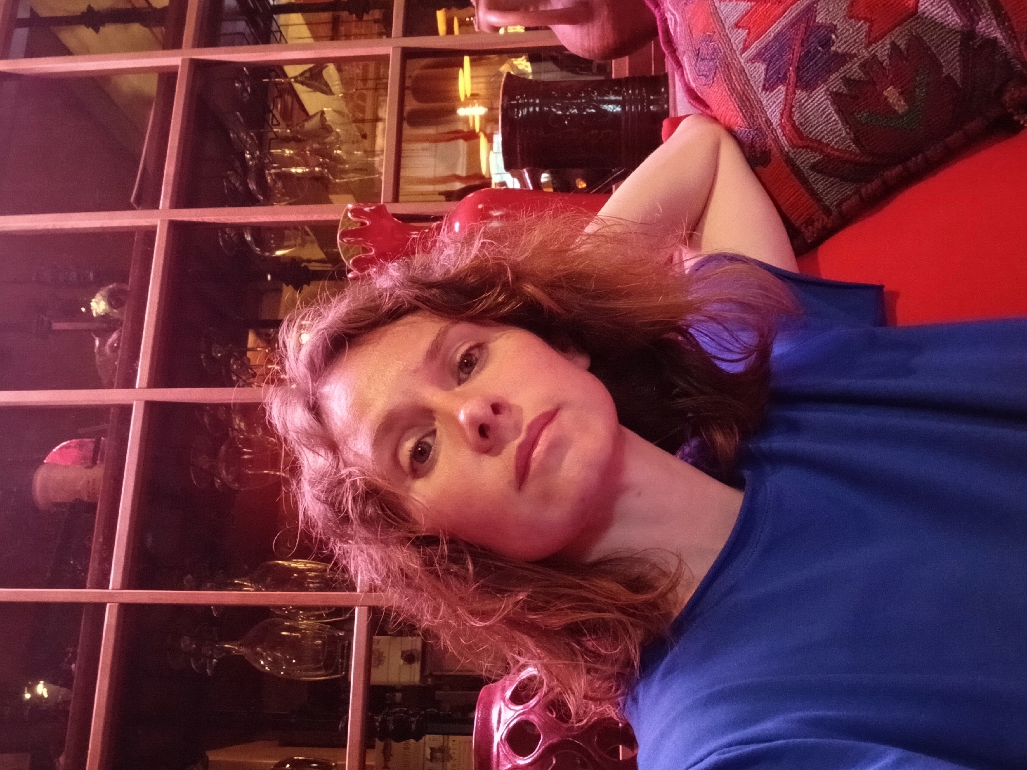 A young woman with curly red hair is taking a selfie in a dimly lit room with a reddish hue. She is wearing a bright blue t-shirt and is resting her right arm on a patterned cushion. Behind her, a wooden shelving unit is filled with various glass bottles, jars, and decorative items. The shelving unit appears to be part of the interior decor of a bar or restaurant. There are no visible external environmental cues to determine the location in Nizhny Novgorod, Russia.