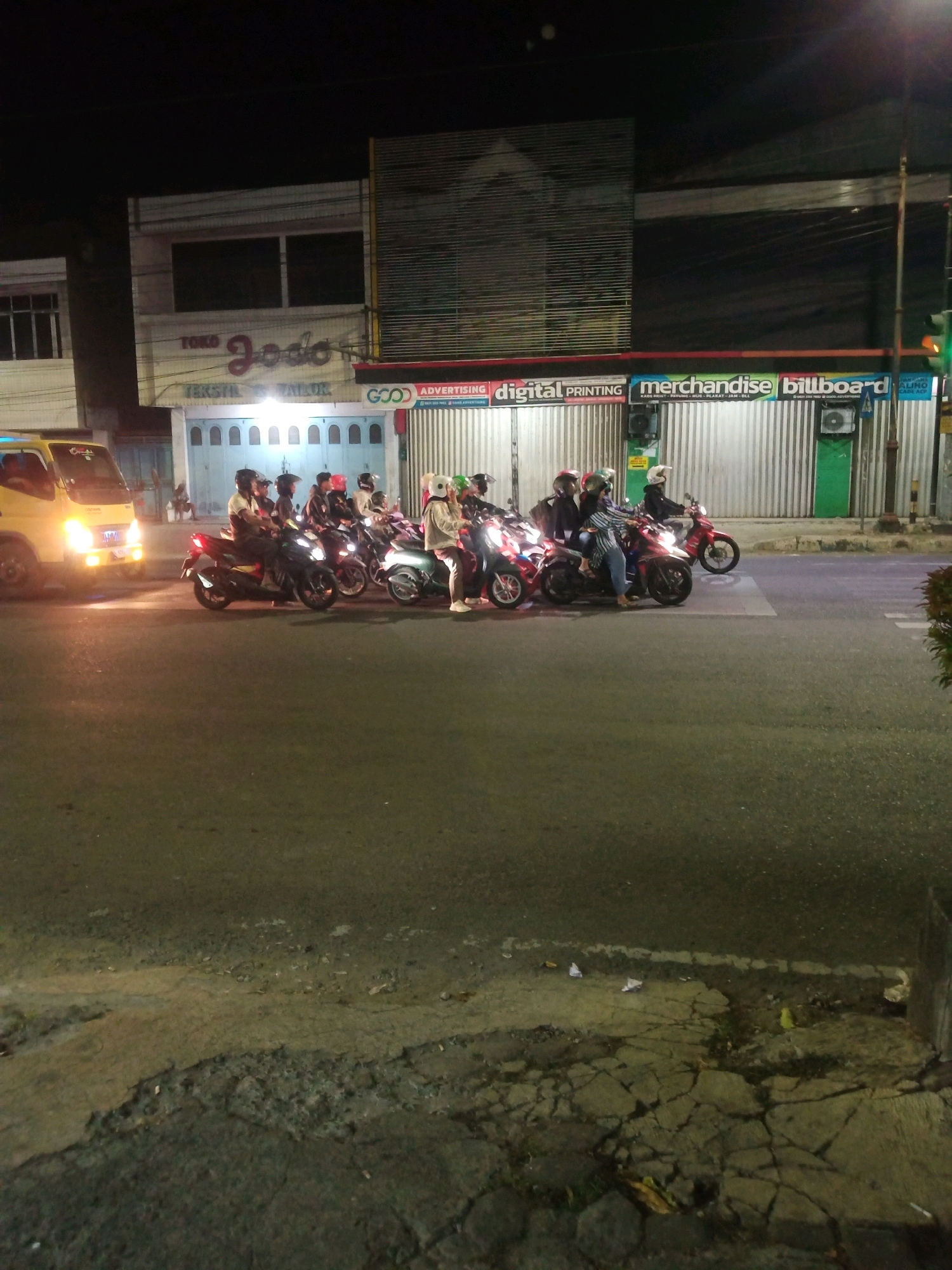 At night, a group of people on motorcycles are stopped at an intersection in Purwokerto, Indonesia. The street is paved and appears to be well-lit by streetlights and the headlights of the vehicles. The buildings lining the street are a mix of commercial establishments, one of which has a sign that reads "TOKO JODO TEKSTIL & TAILOR." Another sign advertises "GOOD ADVERTISING DIGITAL PRINTING MERCHANDISE BILLBOARD." The people on the motorcycles are wearing helmets, and some appear to be engaged in conversation while waiting. The overall atmosphere is one of urban nighttime activity.