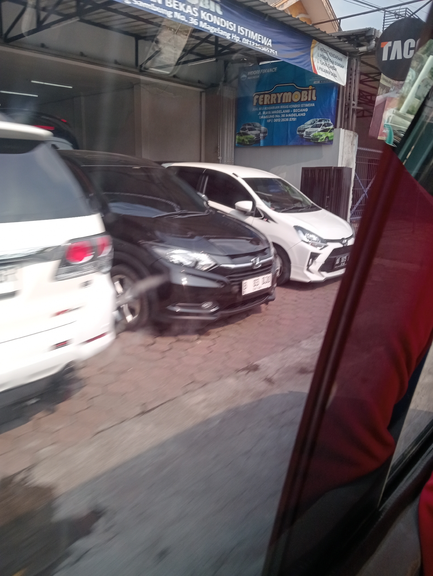 The image depicts a street scene with several cars parked outside a business. A white SUV is partially visible on the left, followed by a black sedan and a white hatchback. A large banner with the text "FERRYMOBIL" and images of cars is displayed on the right side of the frame, indicating a car dealership or service. Other signs and structures suggest a commercial area. The overall impression is of a sunlit outdoor environment, likely in a commercial district of Jambewangi, Indonesia, given the text on the banner.